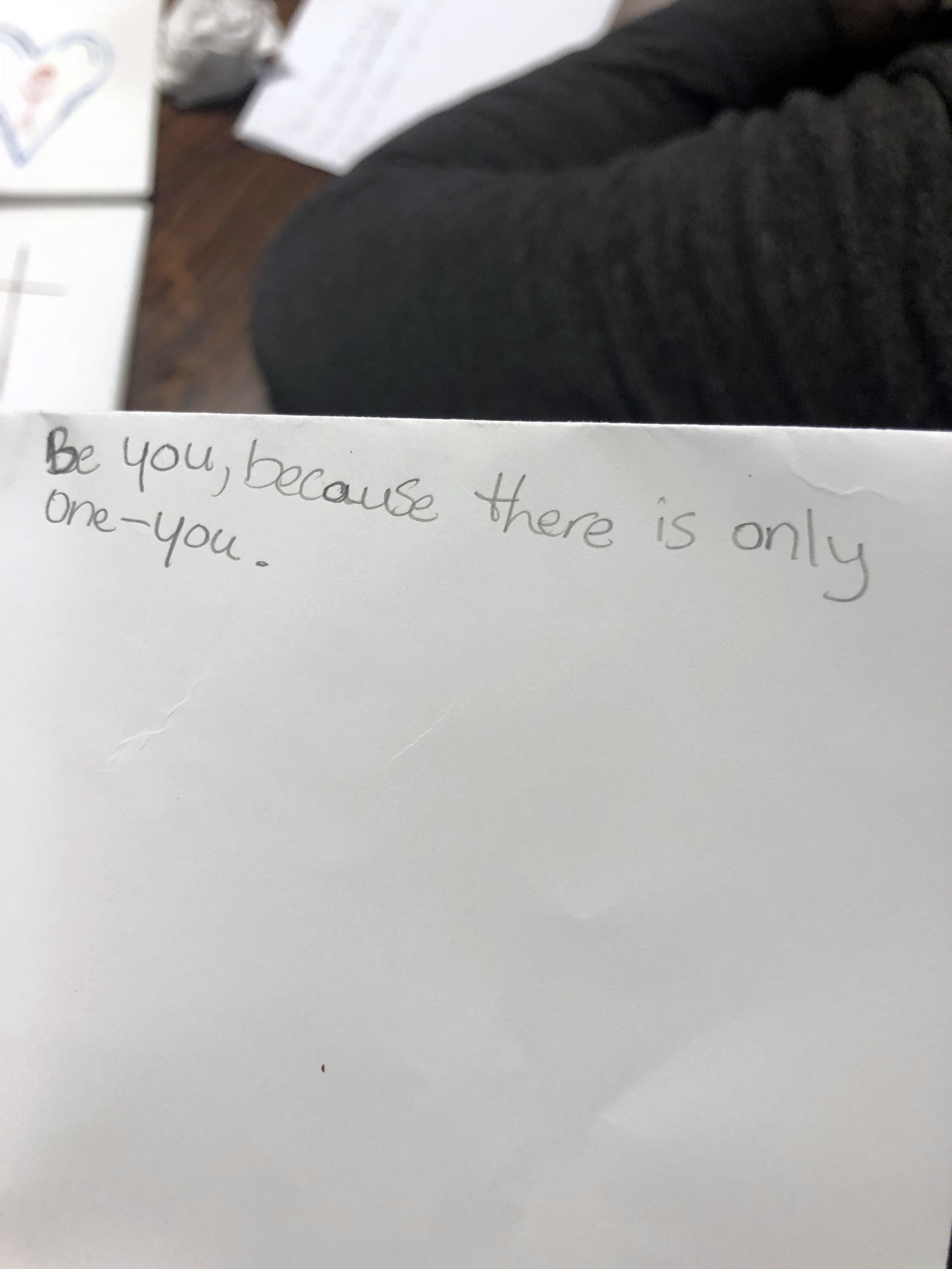 A handwritten note on a white piece of paper that says, 'Be you, because there is only one-you.' The background shows part of a person's dark clothing and some papers on a wooden surface.