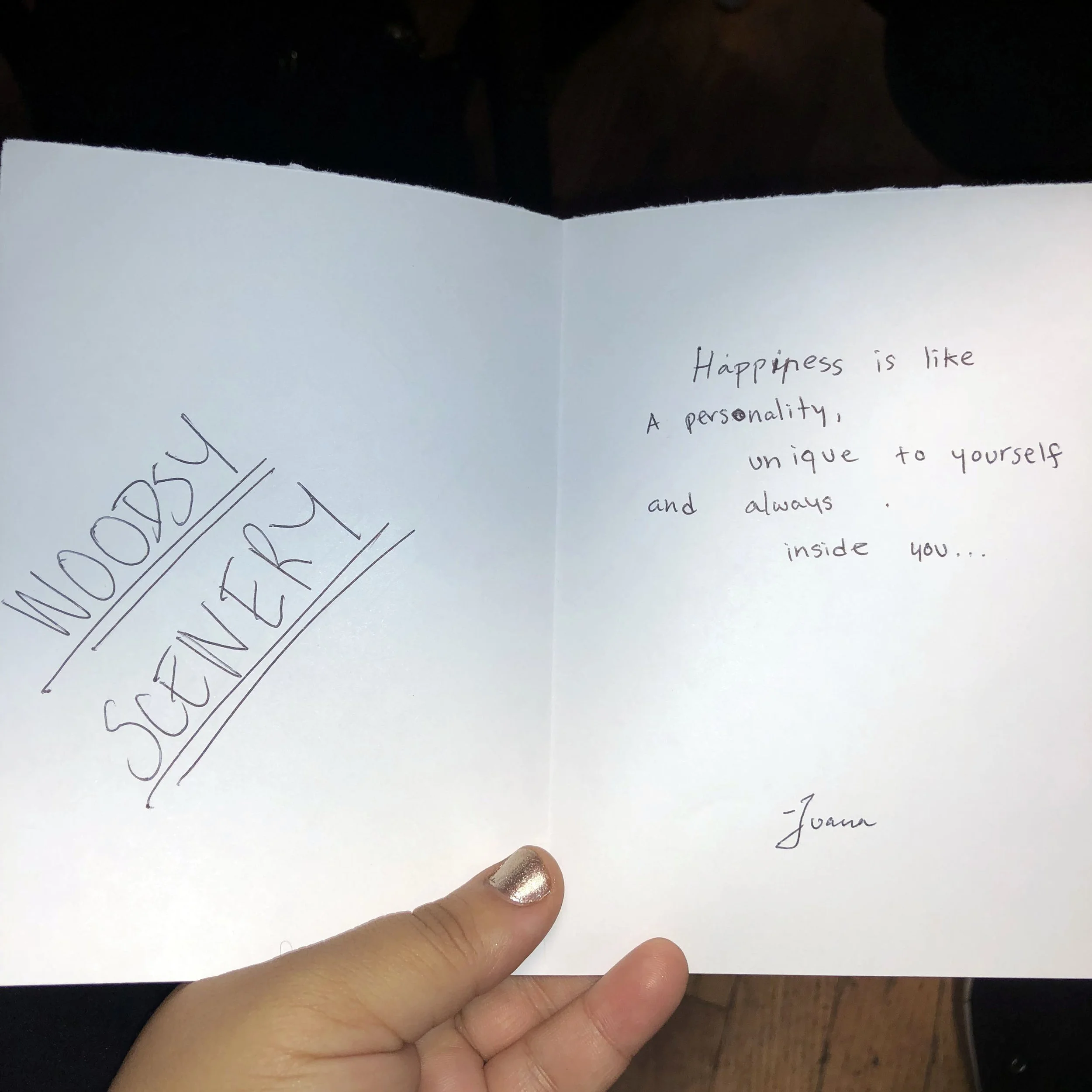 A handwritten note in a card titled 'Words of Sanctuary' with a quote about happiness, inside a person's hand with a metallic nail polish, on a wooden surface.