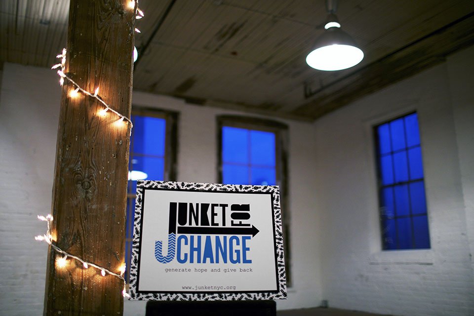 A sign on a table reads 'Junket to Change' with a website link and the slogan 'generate hope and give back'. The background shows a rustic interior with brick walls, large windows, and a wooden ceiling, with string lights wrapped around a wooden post.