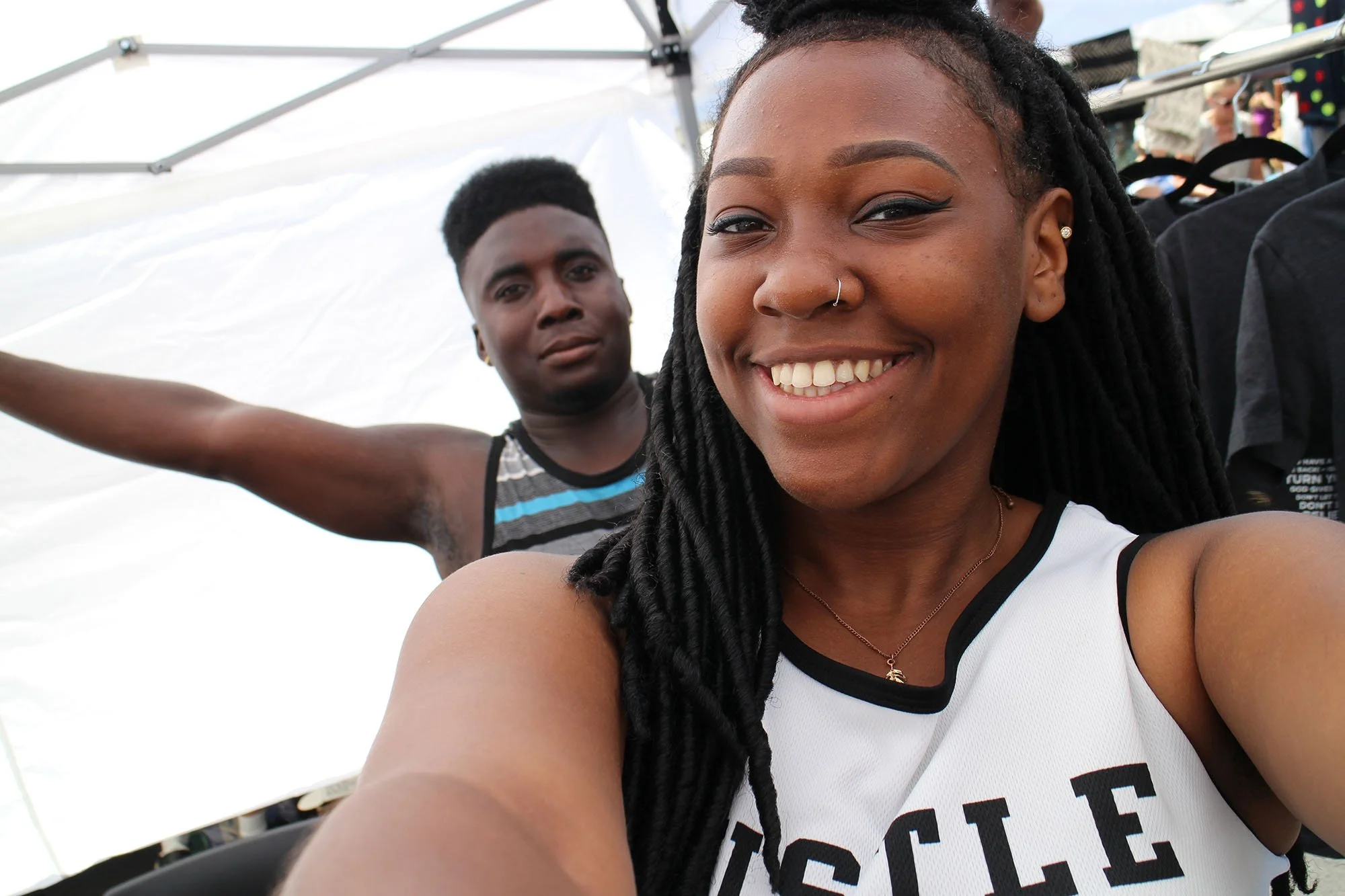 Two young adults taking a selfie at an outdoor event. The woman in the foreground has long black dreadlocks, a nose ring, and is wearing a white sports jersey that says 'CYCLE.' The man behind her has short black hair, a neutral expression, and is wearing a sleeveless shirt with black, white, and gray stripes.