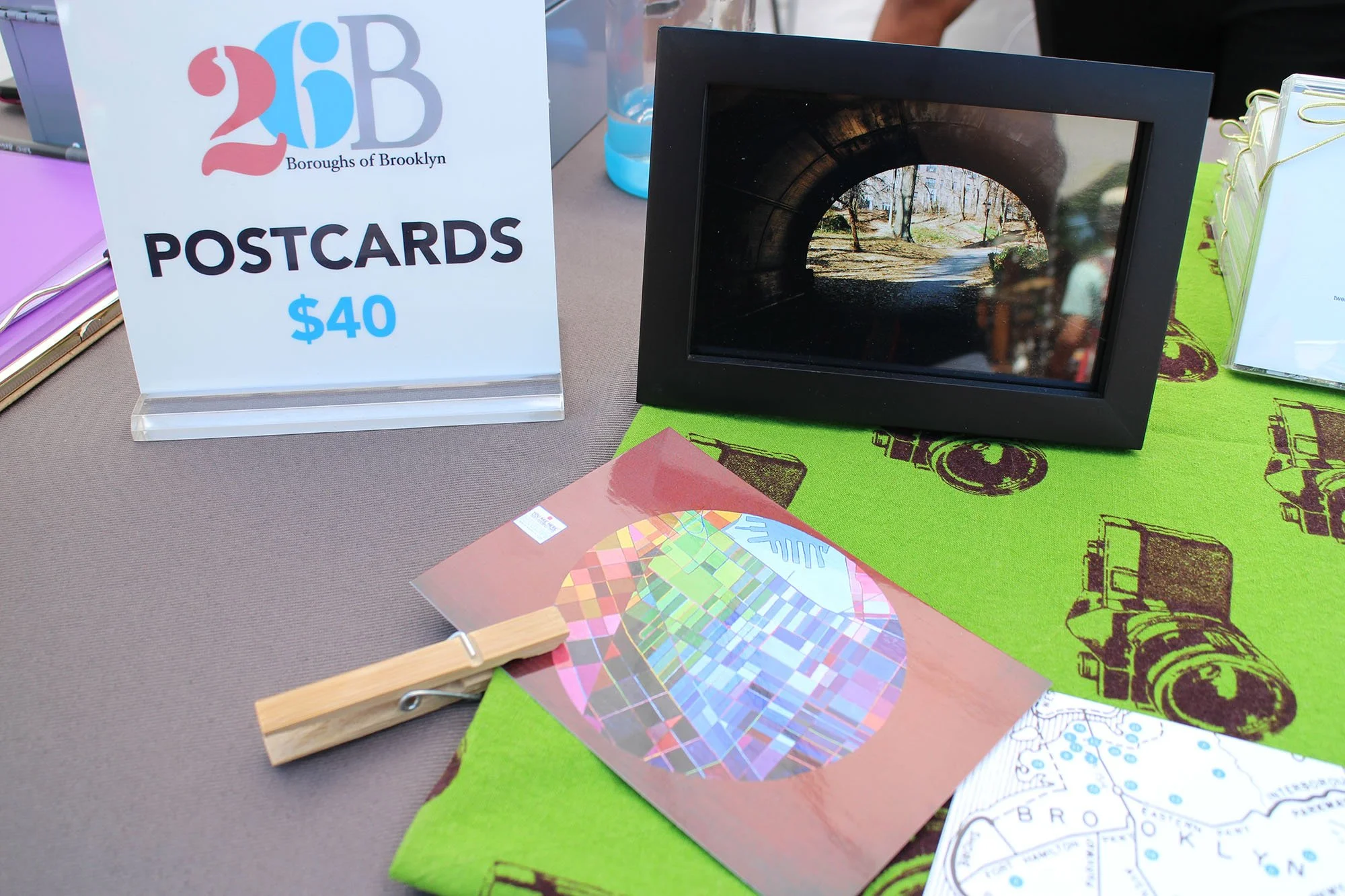 A table display of Brooklyn Borough postcards priced at $40, a framed photograph, a color wheel, and a map with a green cloth featuring camera illustrations, at an outdoor event.