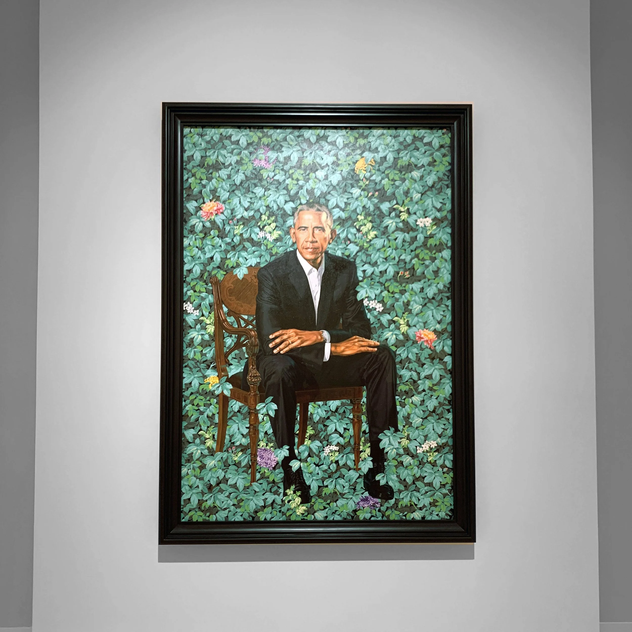 Portrait of Barack Obama sitting on a wooden chair surrounded by lush green foliage and colorful flowers.