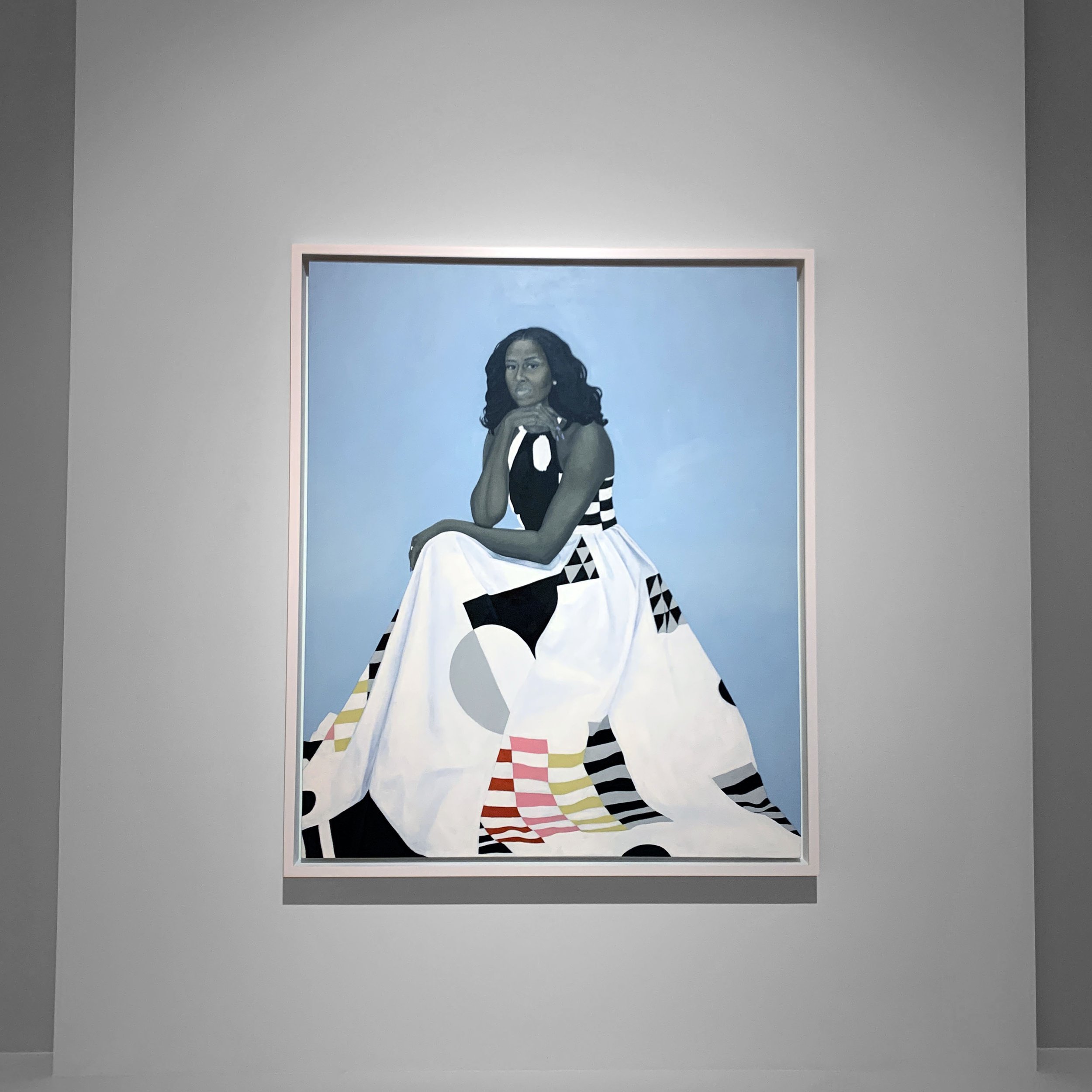 Portrait of a woman with dark curly hair, seated with her chin resting on her hand, wearing a black and white patterned dress against a light blue background in a gallery setting.