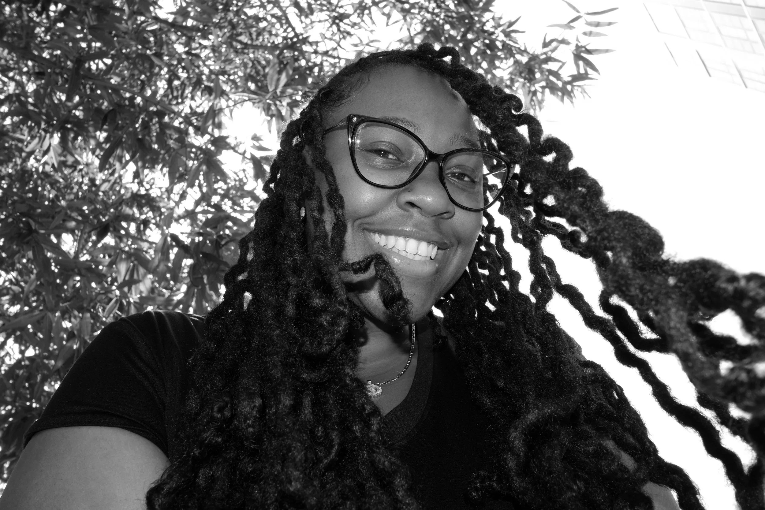 A smiling woman with glasses and dreadlocks wearing a dark sweater with a white V-neck and a black turtleneck underneath, sitting in front of a white brick wall decorated with a hanging garland of leaves.