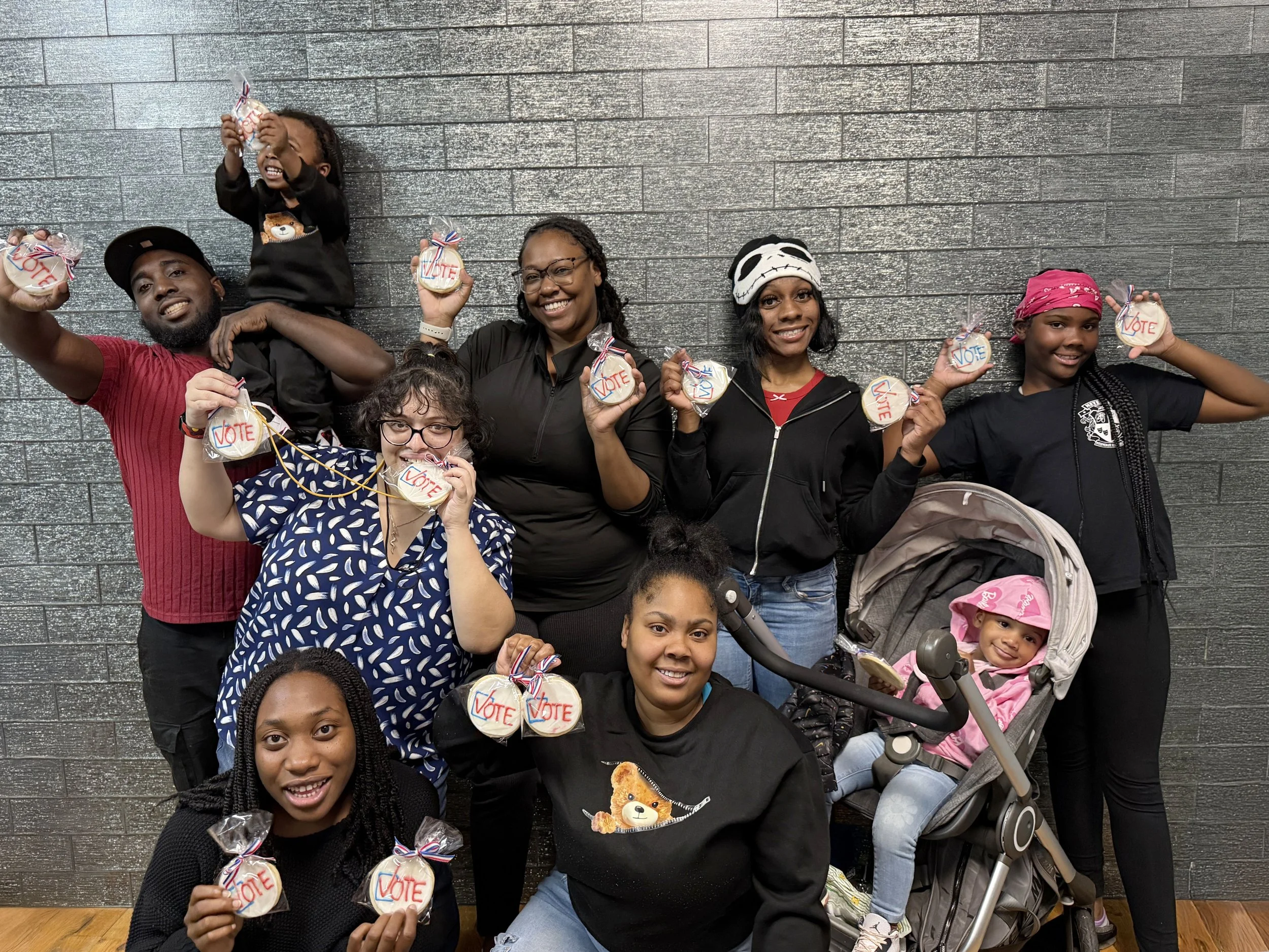Group of seven young people posing together against a gray brick wall, some standing and some kneeling, smiling and making playful gestures.