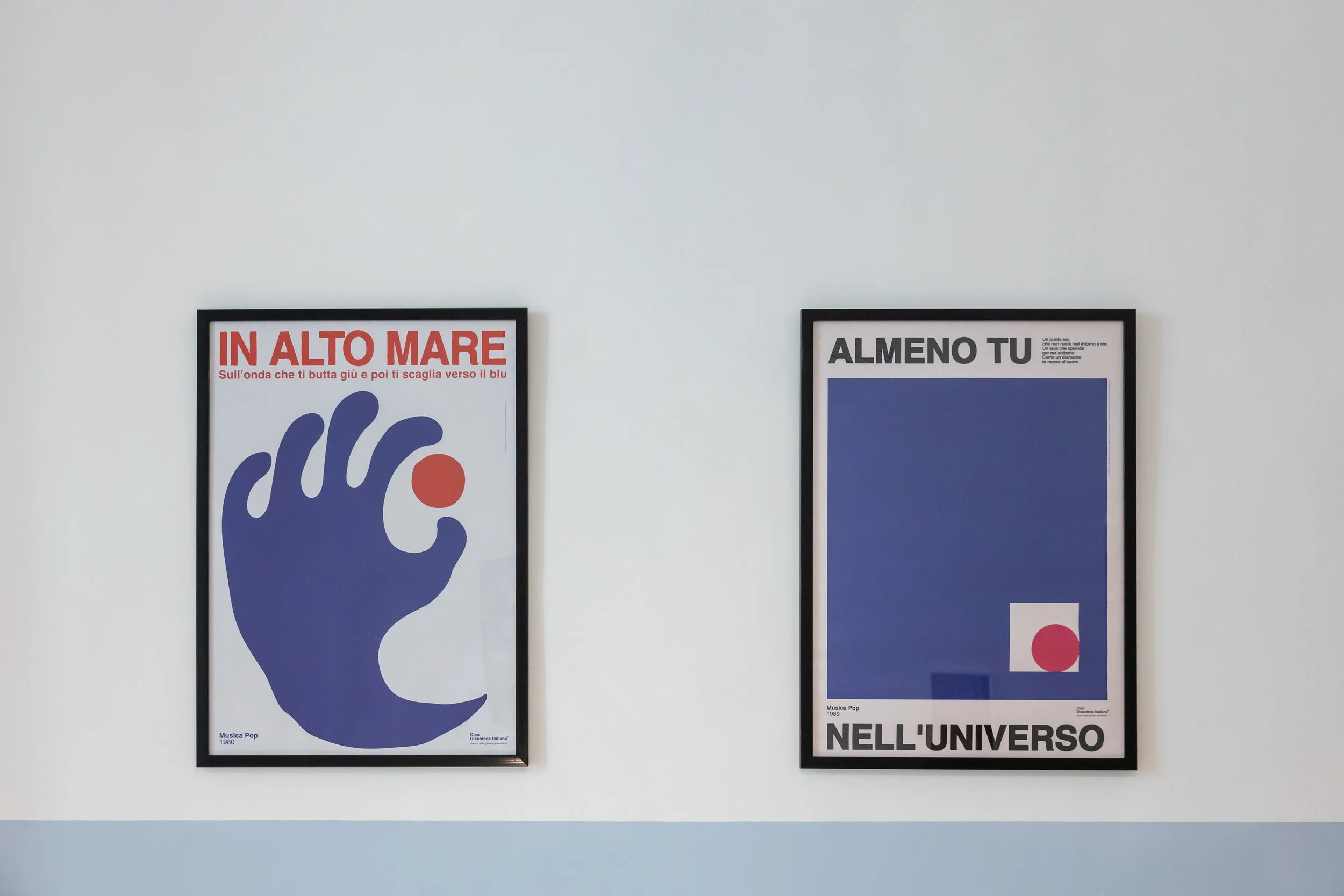 Two framed posters on a white wall, one with text in Italian and a stylized hand with a red circle, the other with a blue square and a small red circle.