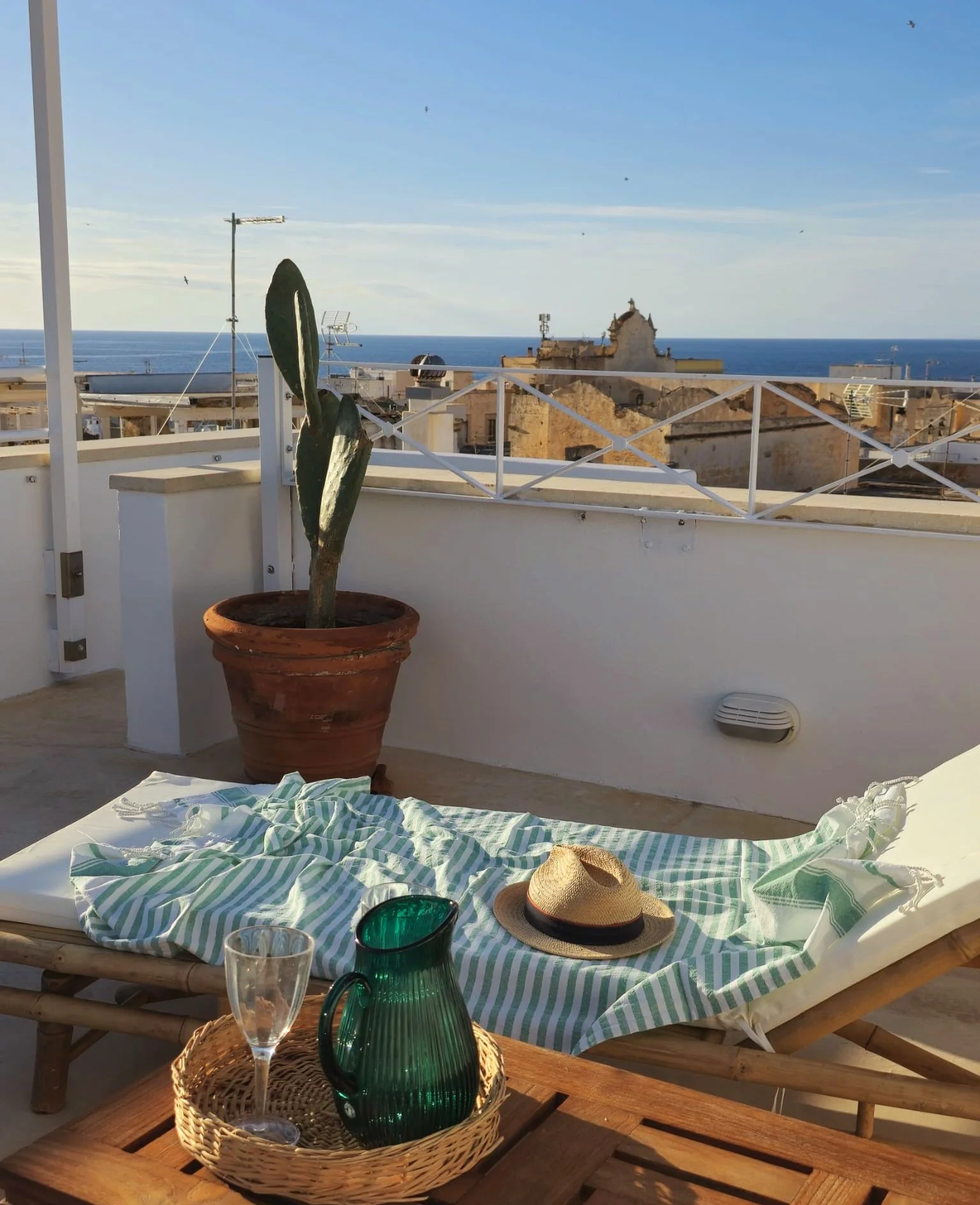 Rooftop terrace with a potted cactus, a sun lounger with a hat, a striped cloth, a green glass pitcher, a clear glass, and a view of the sea and city buildings in the background.