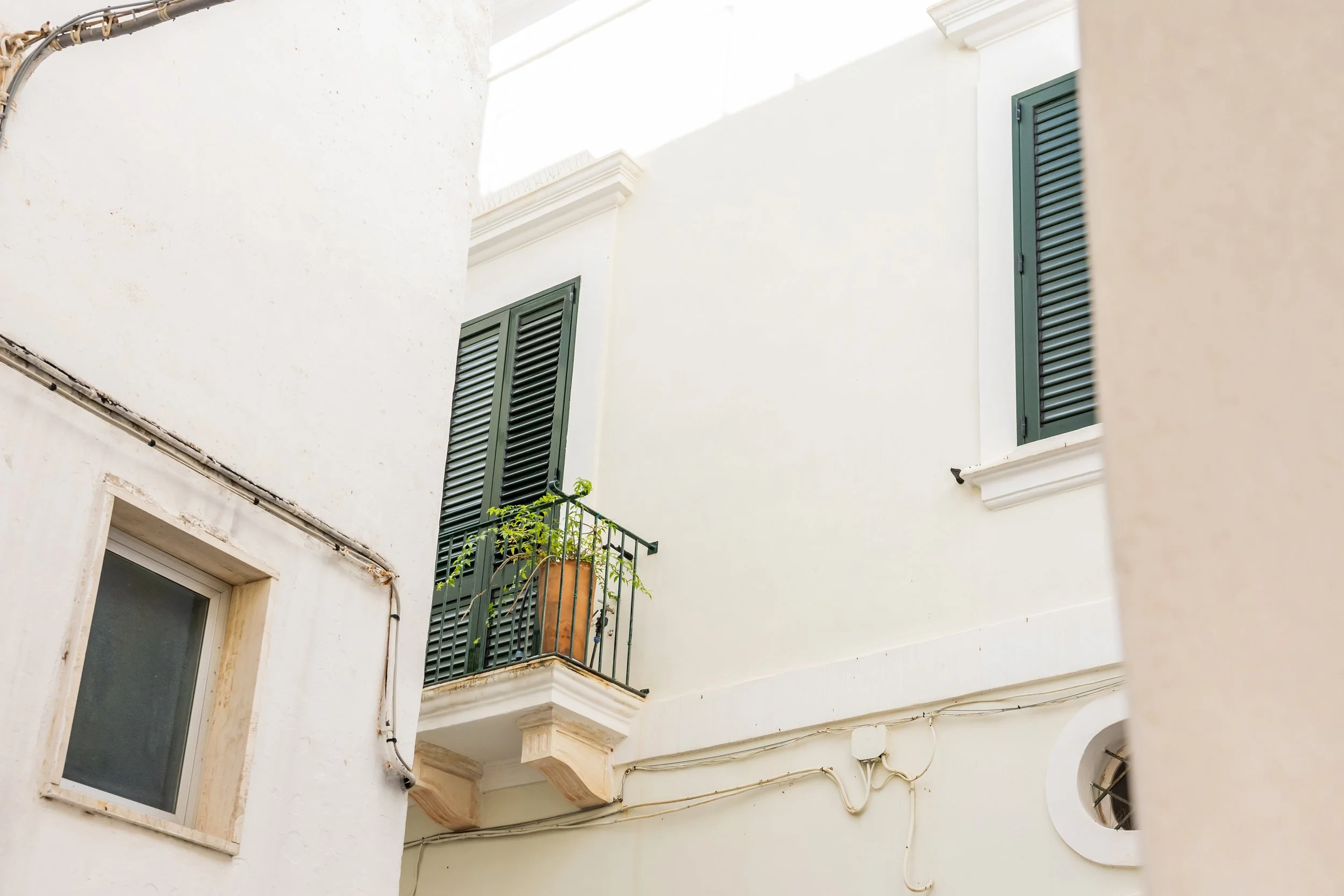 White building with green window shutters and a small balcony with a potted plant, located in a sunny area.