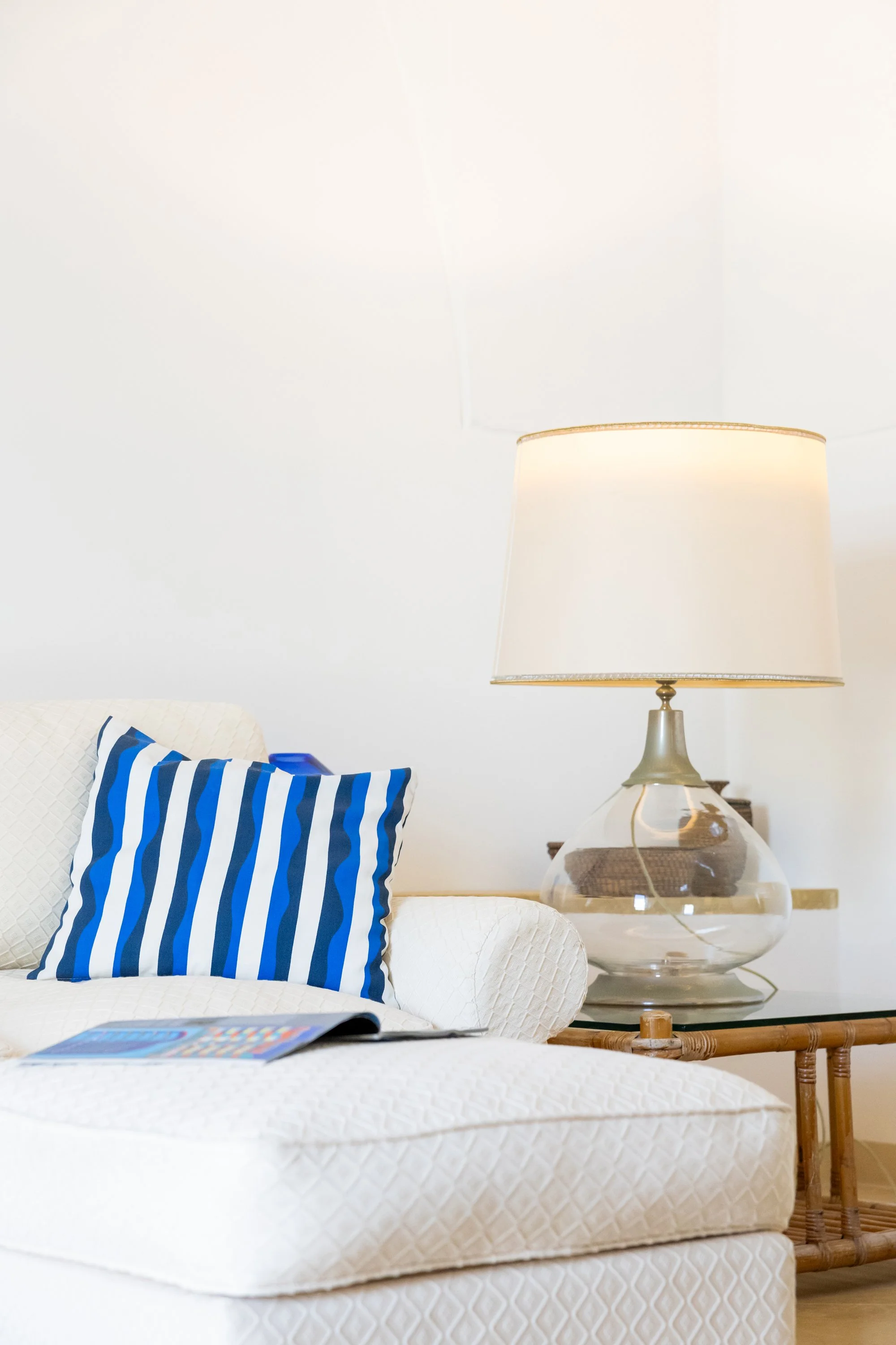 A cozy white tufted sofa with a blue and white striped pillow, a magazine, a glass table with a large glass lamp, and a woven basket on a wooden side table in a bright room.