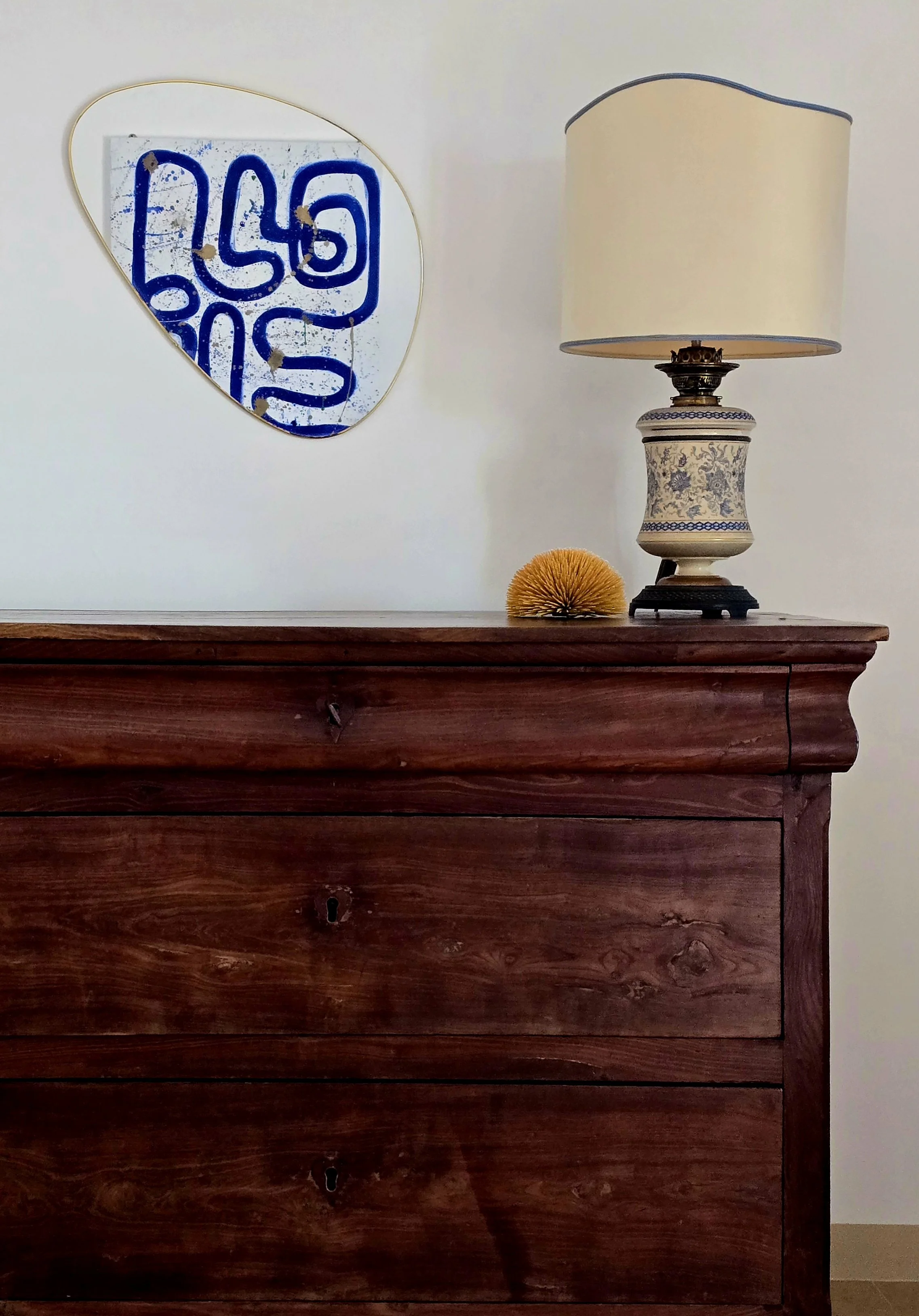 A wooden dresser with a decorative table lamp, a yellow spherical object, and an abstract wall mirror with blue elements hanging on a plain white wall.