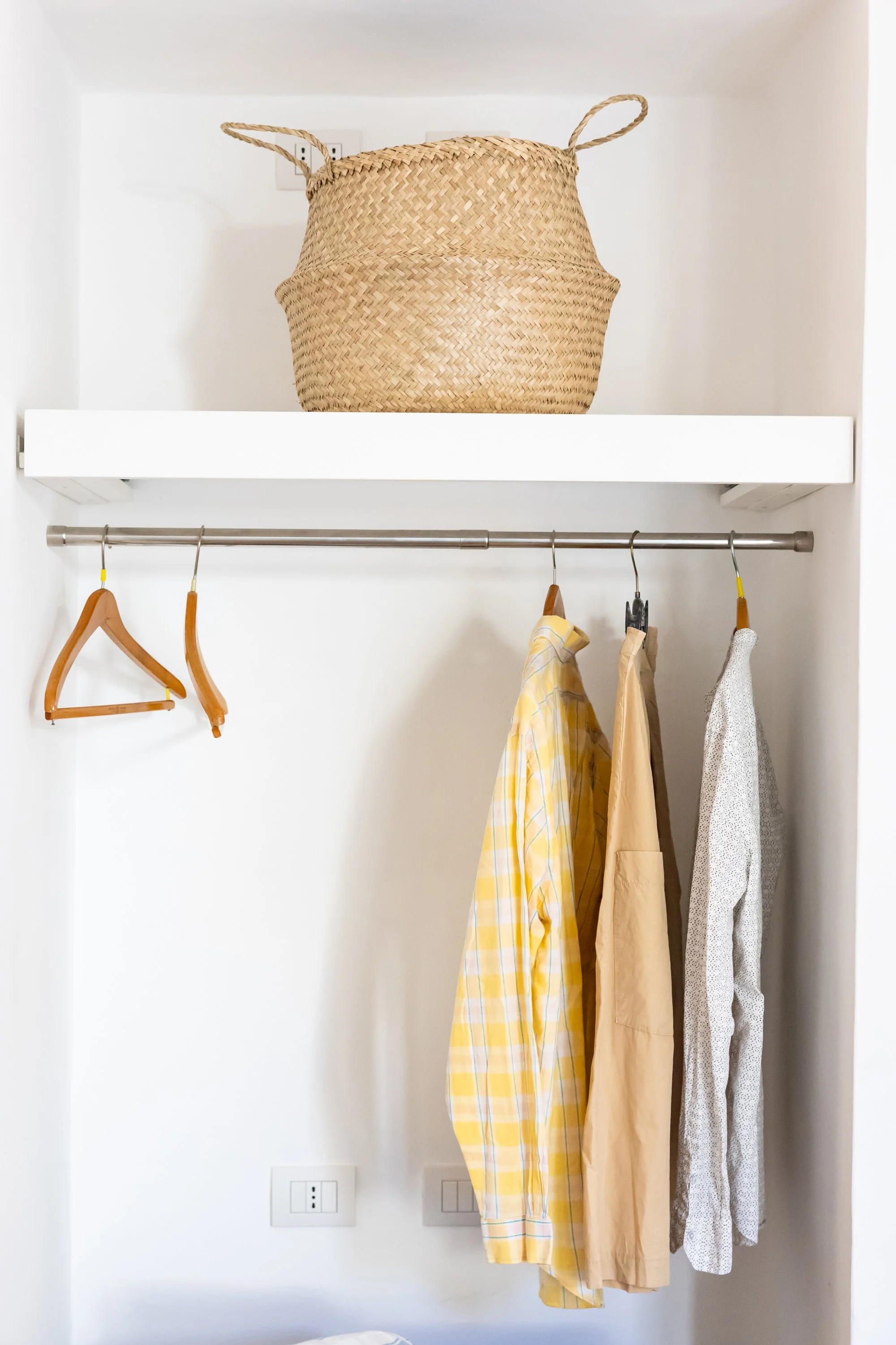 Closet with a woven basket on a white shelf above, and yellow, beige, and gray clothing hanging on a metal rod.
