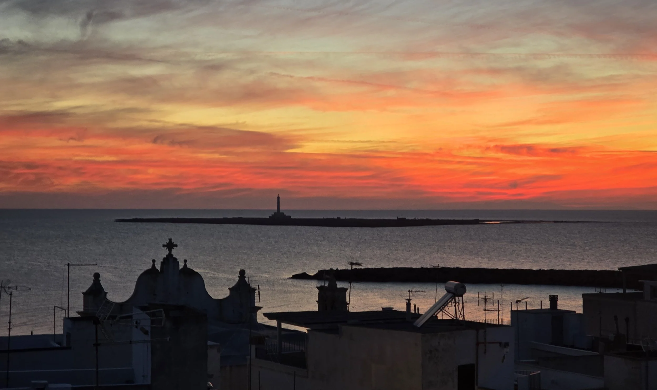 Breathtaking sunset over the sea seen from the rooftop of Palazzo Dolce Vita, with the lighthouse on the horizon and the rooftops of Gallipoli Old Town in the foreground.