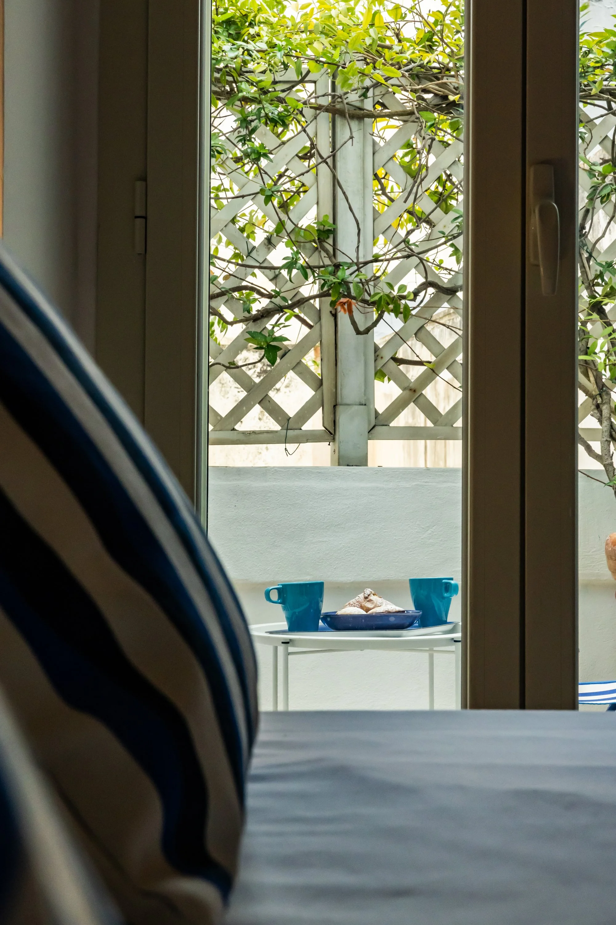 View from inside a room looking out through a window at a small outdoor patio with a white table, two blue cups, a blue plate with seashells, and a background of a lattice fence with green foliage.