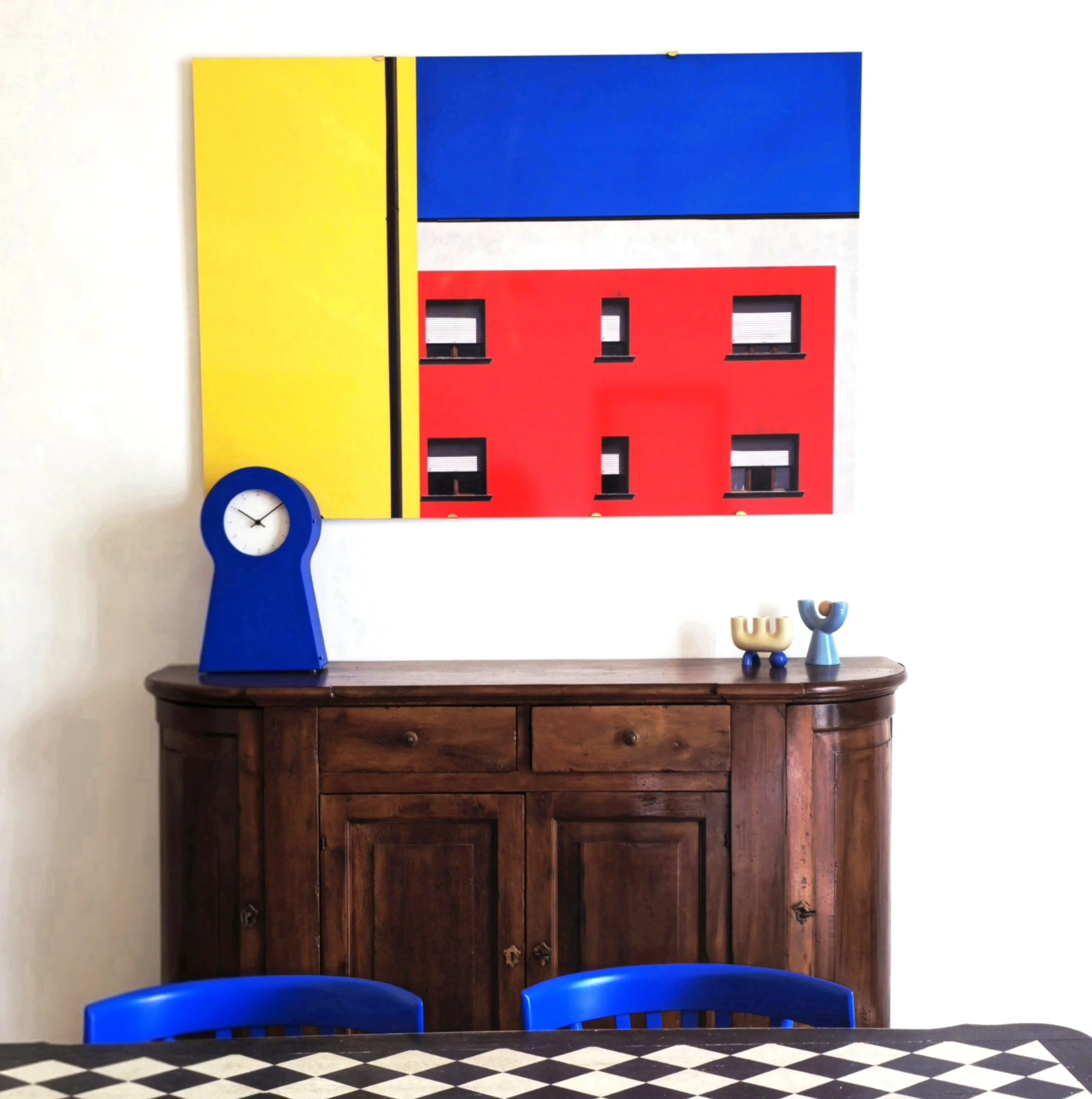 A wall art piece with bold blocks of yellow, blue, white, and red above a wooden sideboard. On the sideboard are a blue clock and decorative vases, with white walls and blue chairs in the foreground.