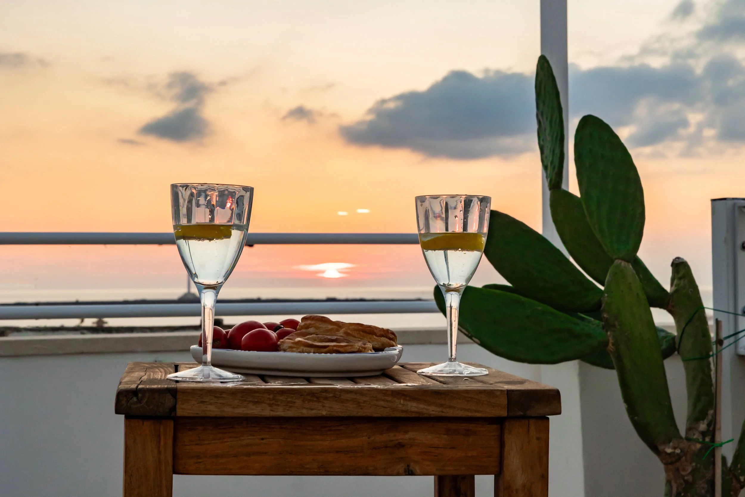 Two glasses of white wine on a wooden table with a plate of cherry tomatoes and pastries, set against a sunset view with a large green plant nearby.