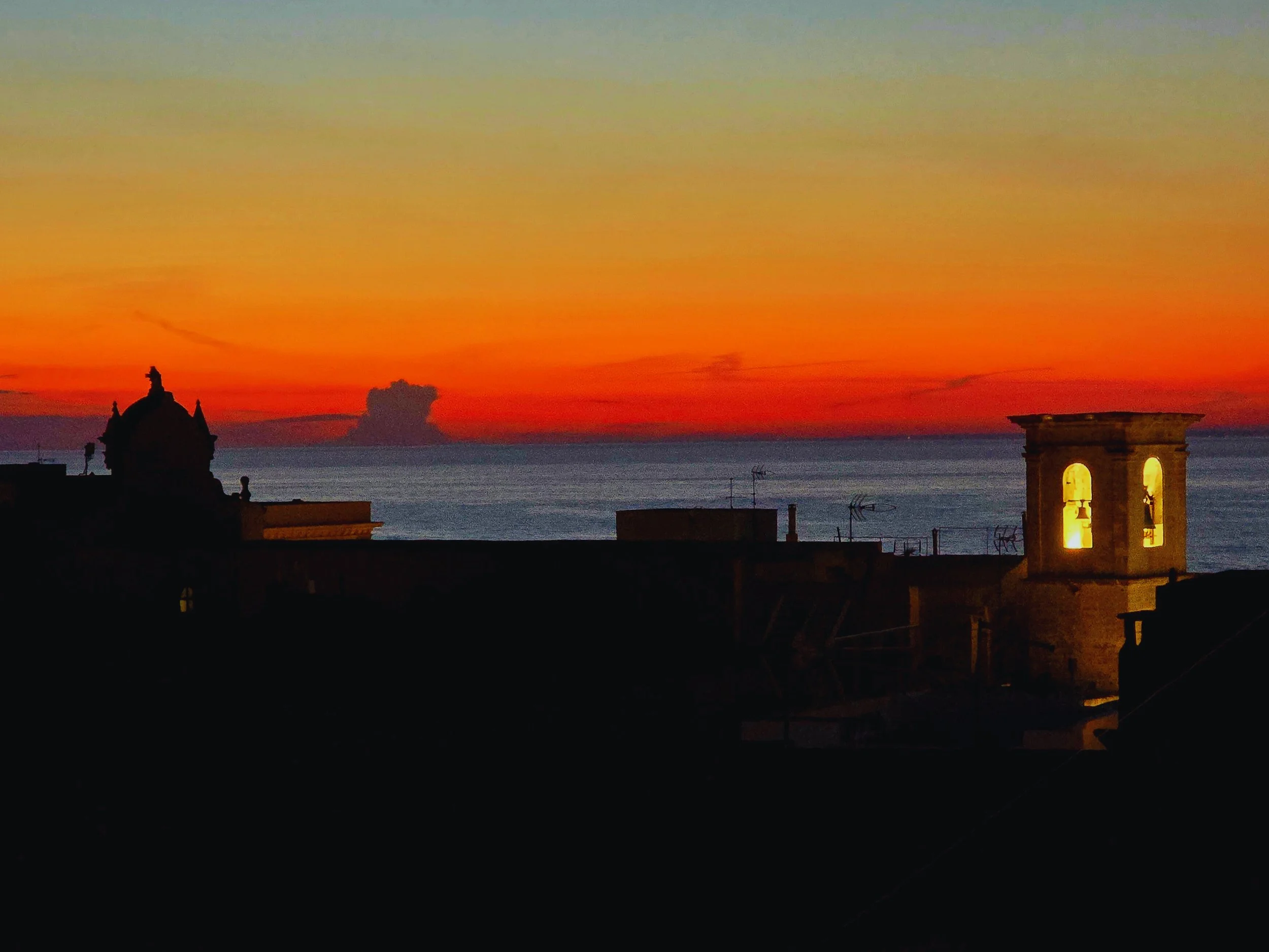 Sunset over the sea seen from the rooftop of Palazzo Dolce Vita, with silhouettes of historic buildings and the illuminated bell tower of Gallipoli Old Town.