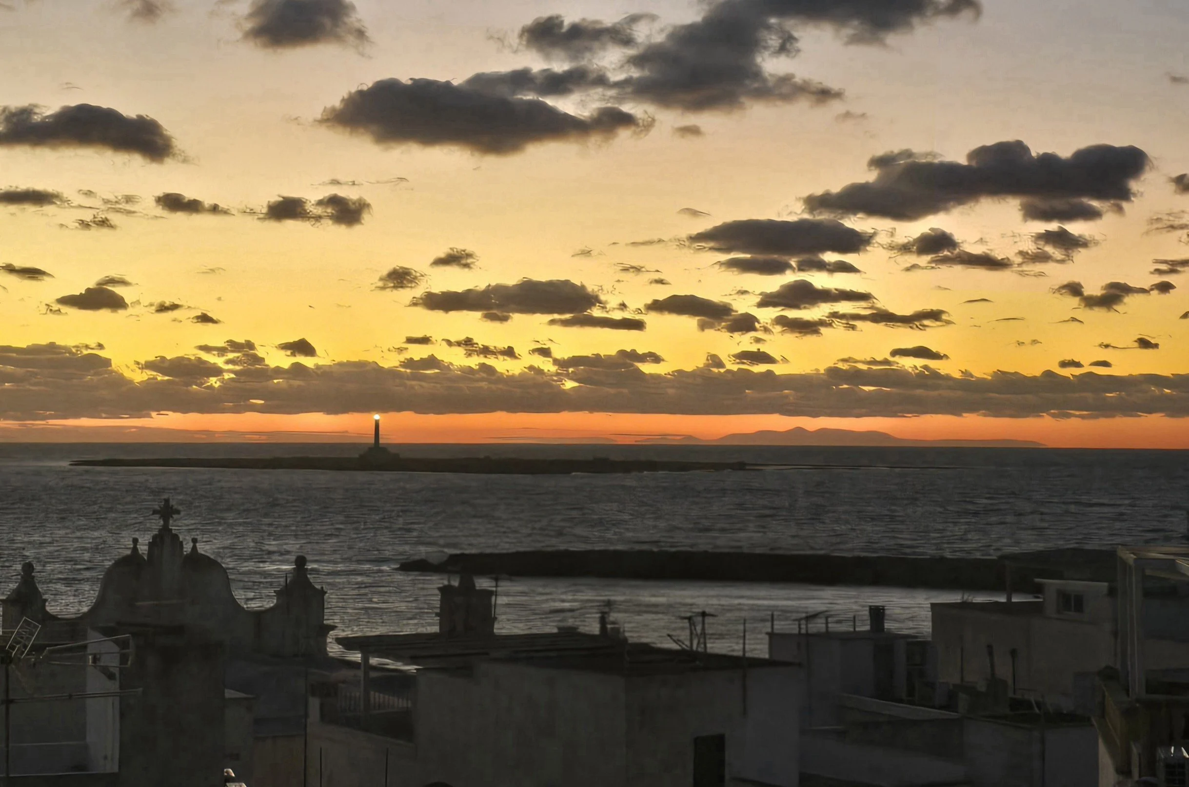 Sunset over the ocean with scattered clouds and a lighthouse on a distant island. Silhouettes of buildings in the foreground.