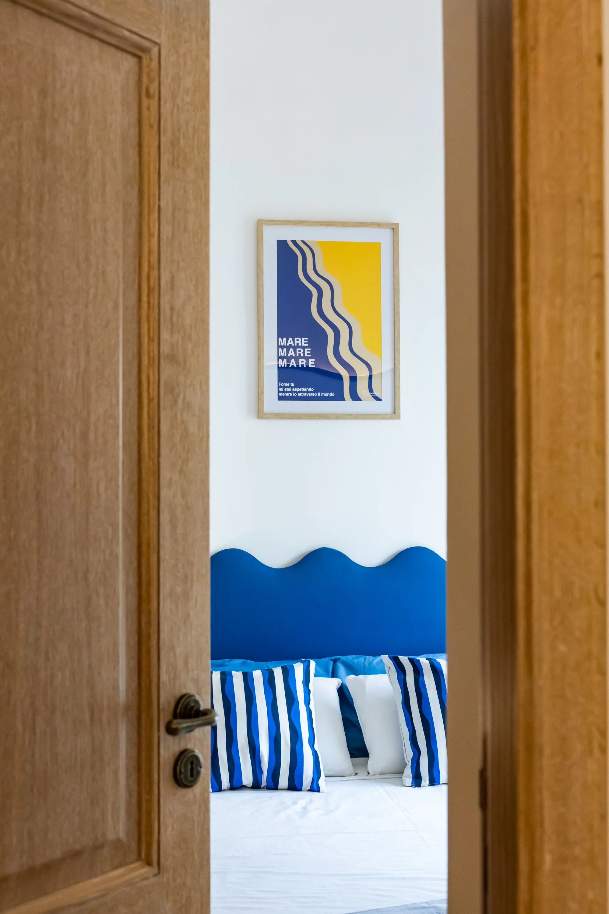 A bedroom seen through an open wooden door. The bed has a blue headboard shaped like waves, with pillows in blue and white stripes, and plain white pillows. There is a framed wall art with wavy lines and the words 'MARE, MARE, MARE' by the wall.