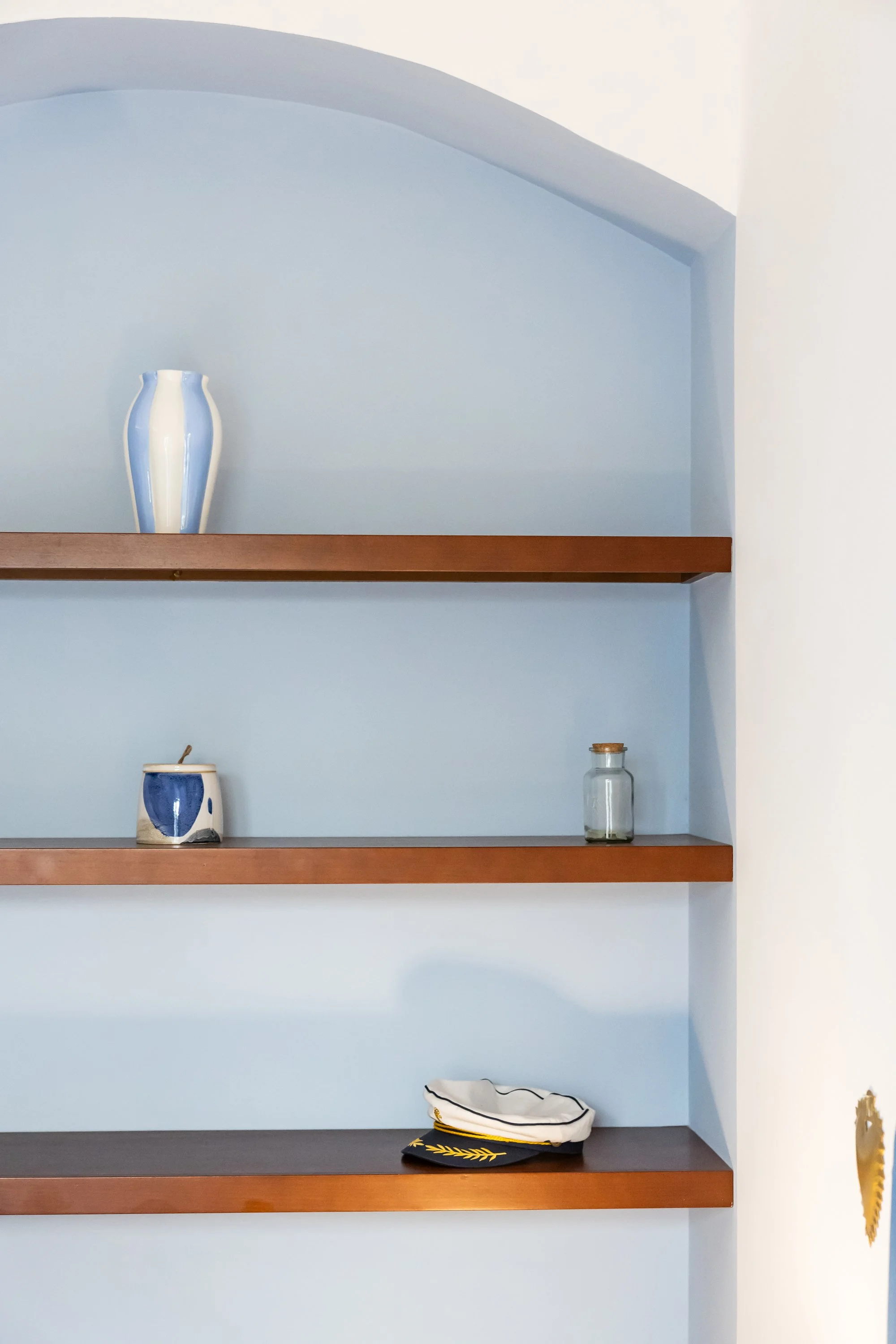 Empty blue wall with three wooden shelves holding decorative items, including a blue and white vase, a small ceramic jar, a glass jar, and a white cap with black and gold embroidery.