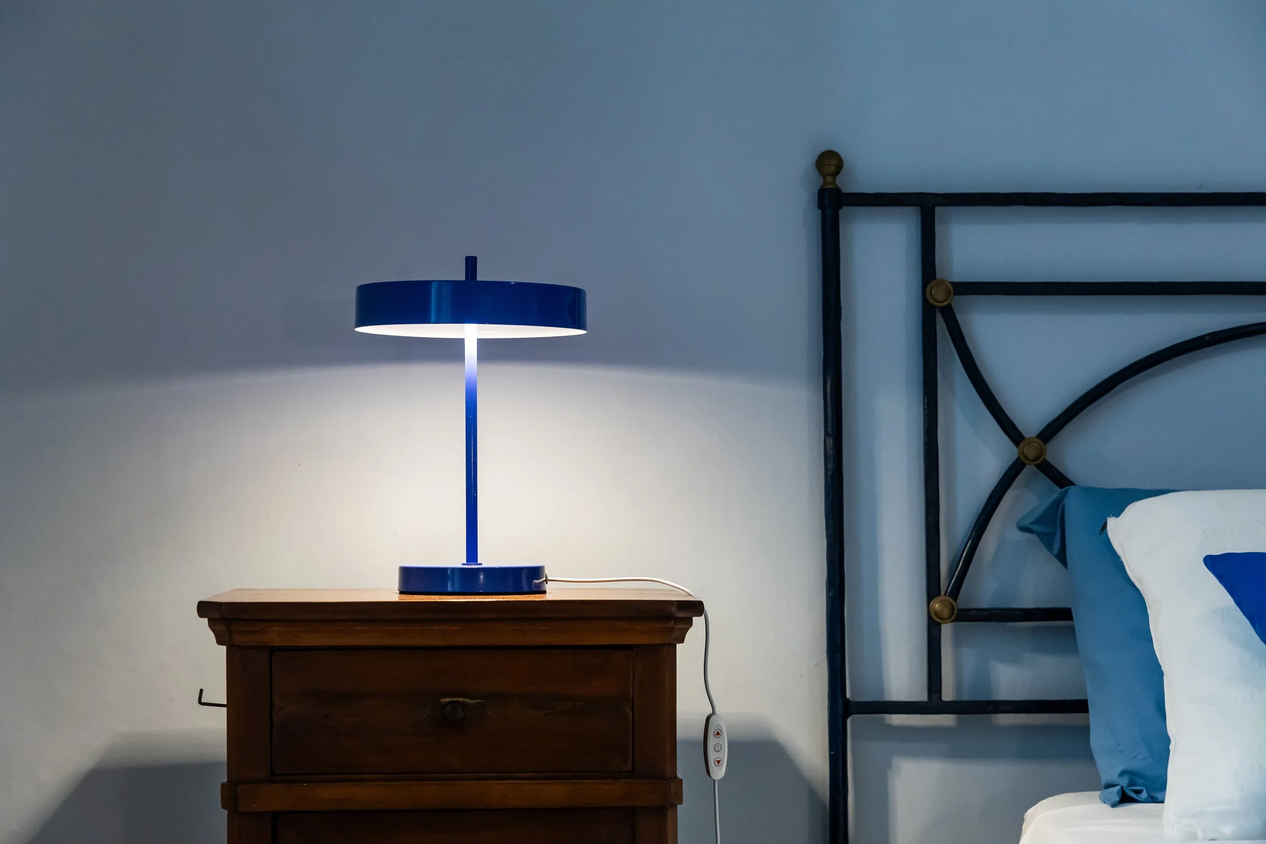 A blue table lamp turned on sits on a wooden nightstand beside a bed with blue and white bedding in a bedroom with white walls.