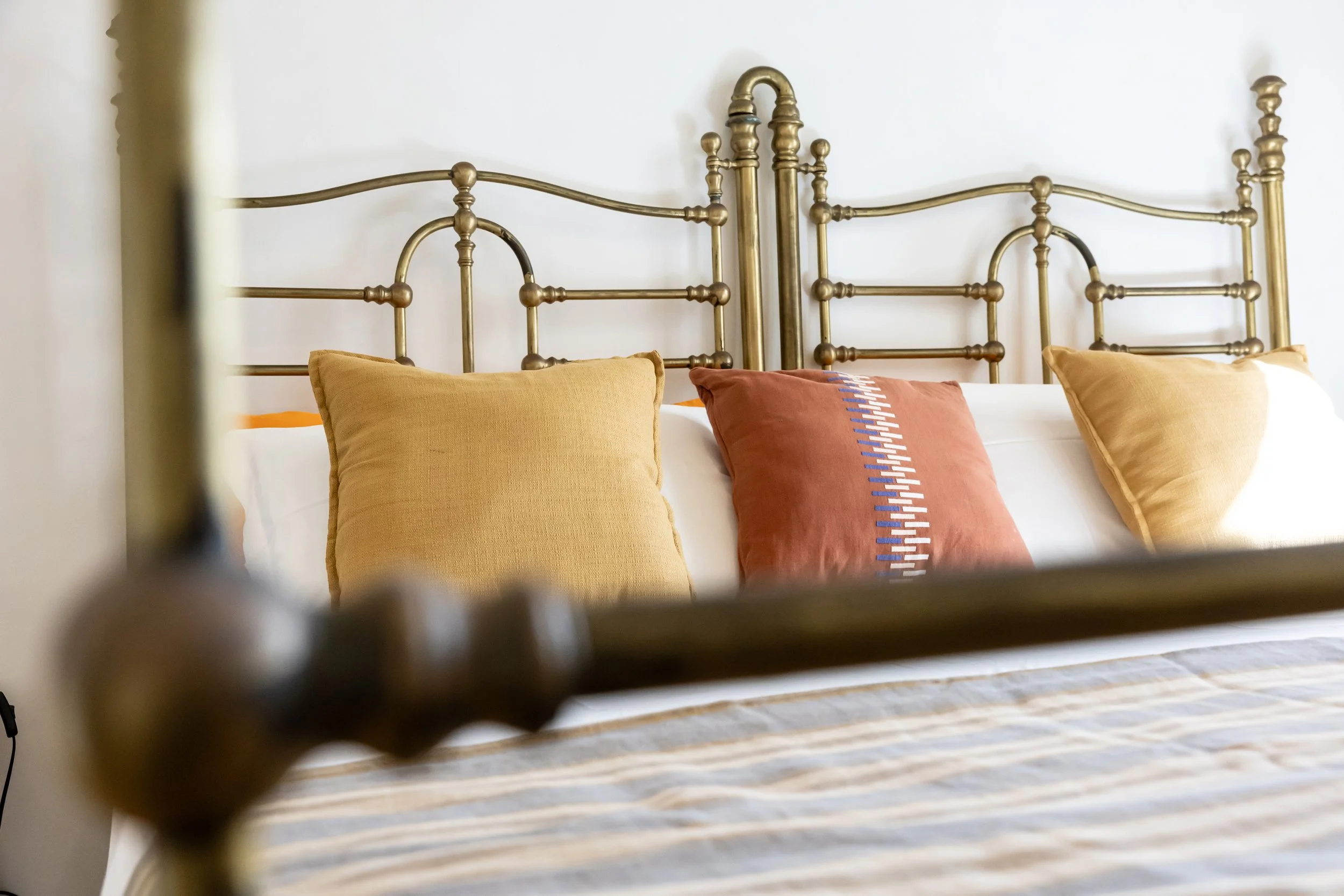 A close-up view of a brass or gold-colored metal bed frame with decorative headboard, with three pillows in yellow, orange with blue and white embroidery, and yellow on a white bed against a plain white wall.
