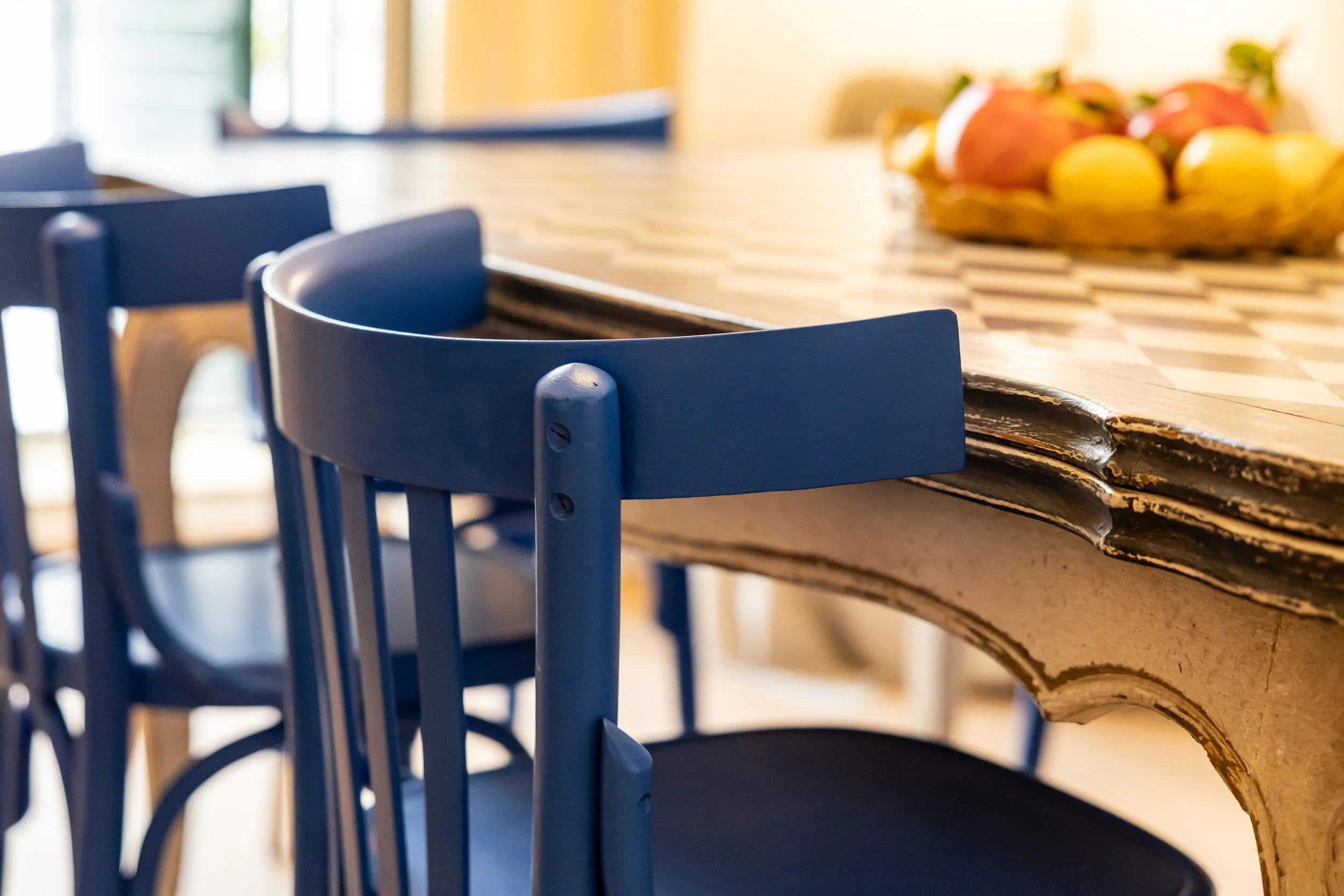Close-up of blue dining chairs around a wooden table with a fruit bowl in the background.