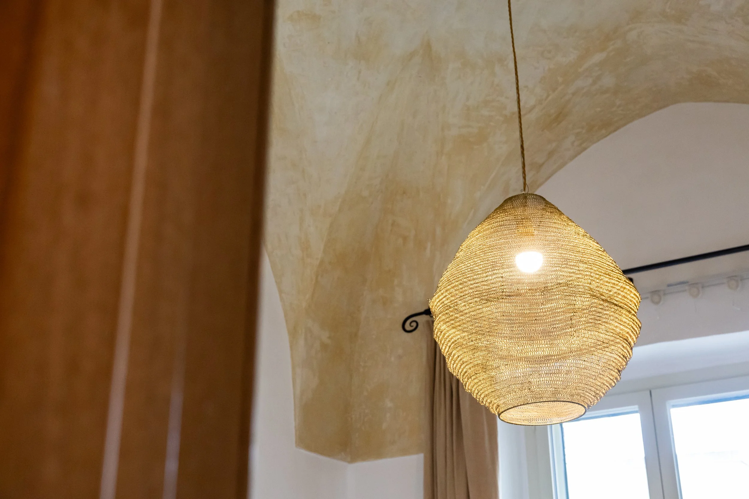 A woven beige pendant light hanging from a ceiling near a window with beige curtains.