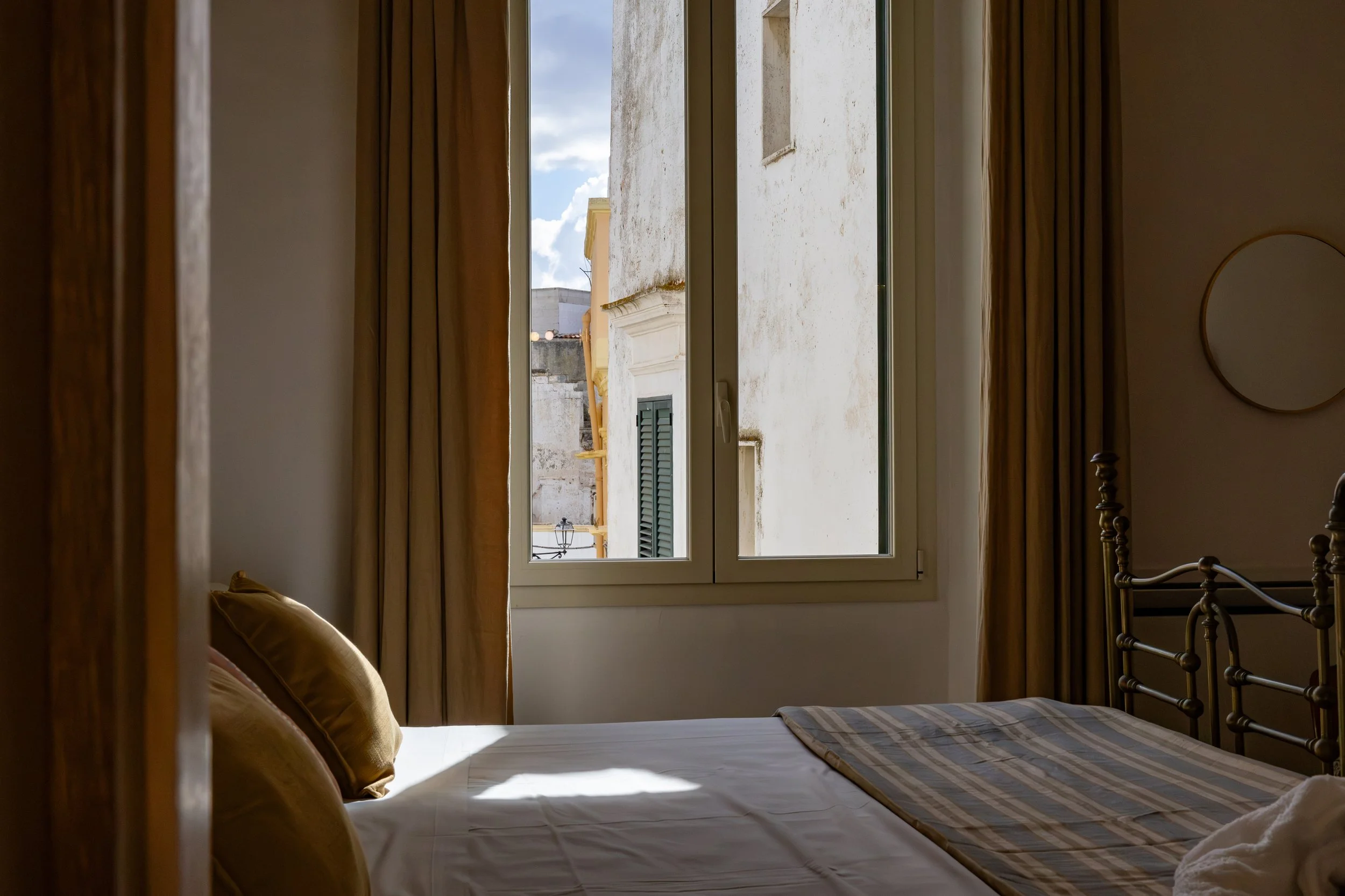 Sunlight illuminates a neatly made bed with pillows in a room with beige curtains, a window showing outside buildings, and partial view of a mirror on the wall.