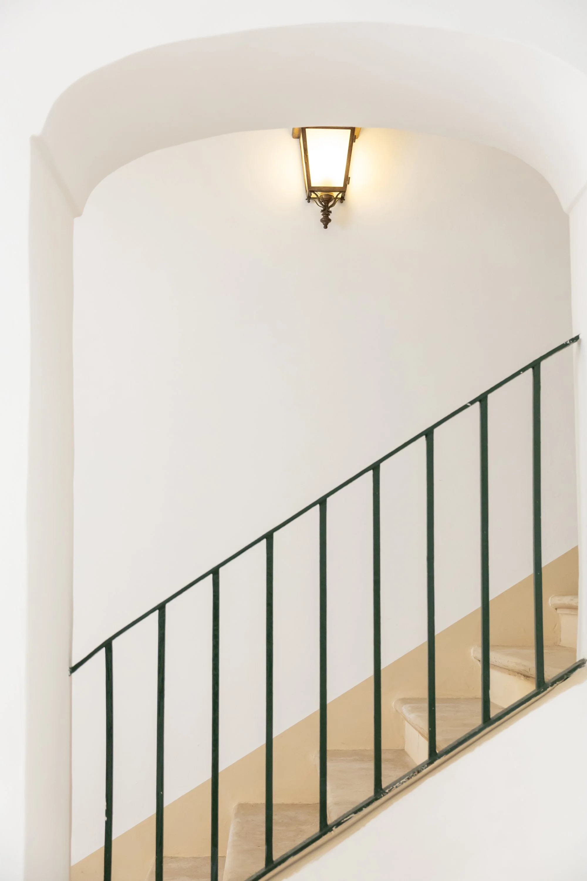 Interior view of a staircase with beige steps, a green railing, white walls, and a ceiling-mounted light fixture.