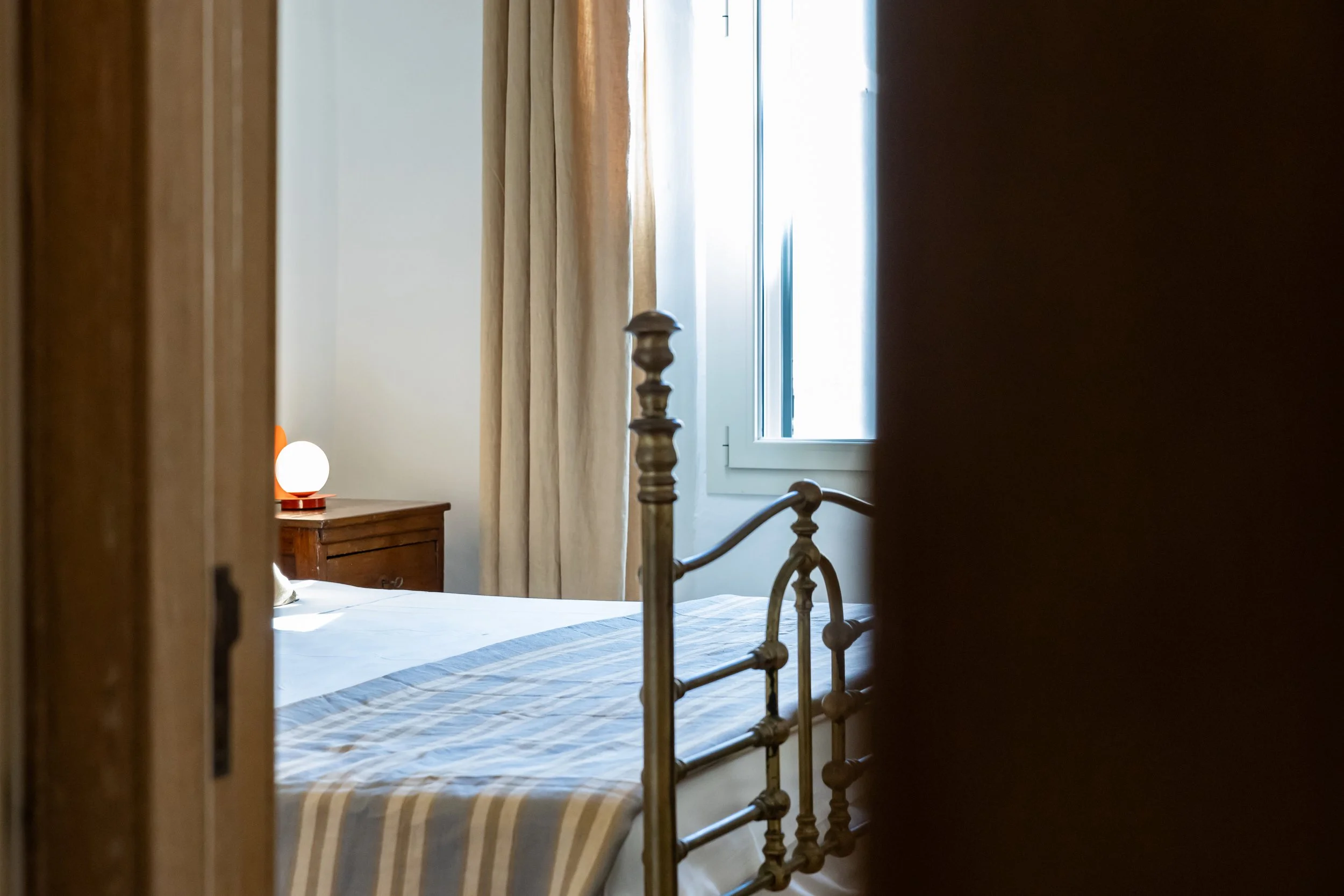 A view of a bedroom through a partially open door, showing a bed with a striped bedspread, a wooden nightstand with a spherical lamp, and a window with beige curtains.