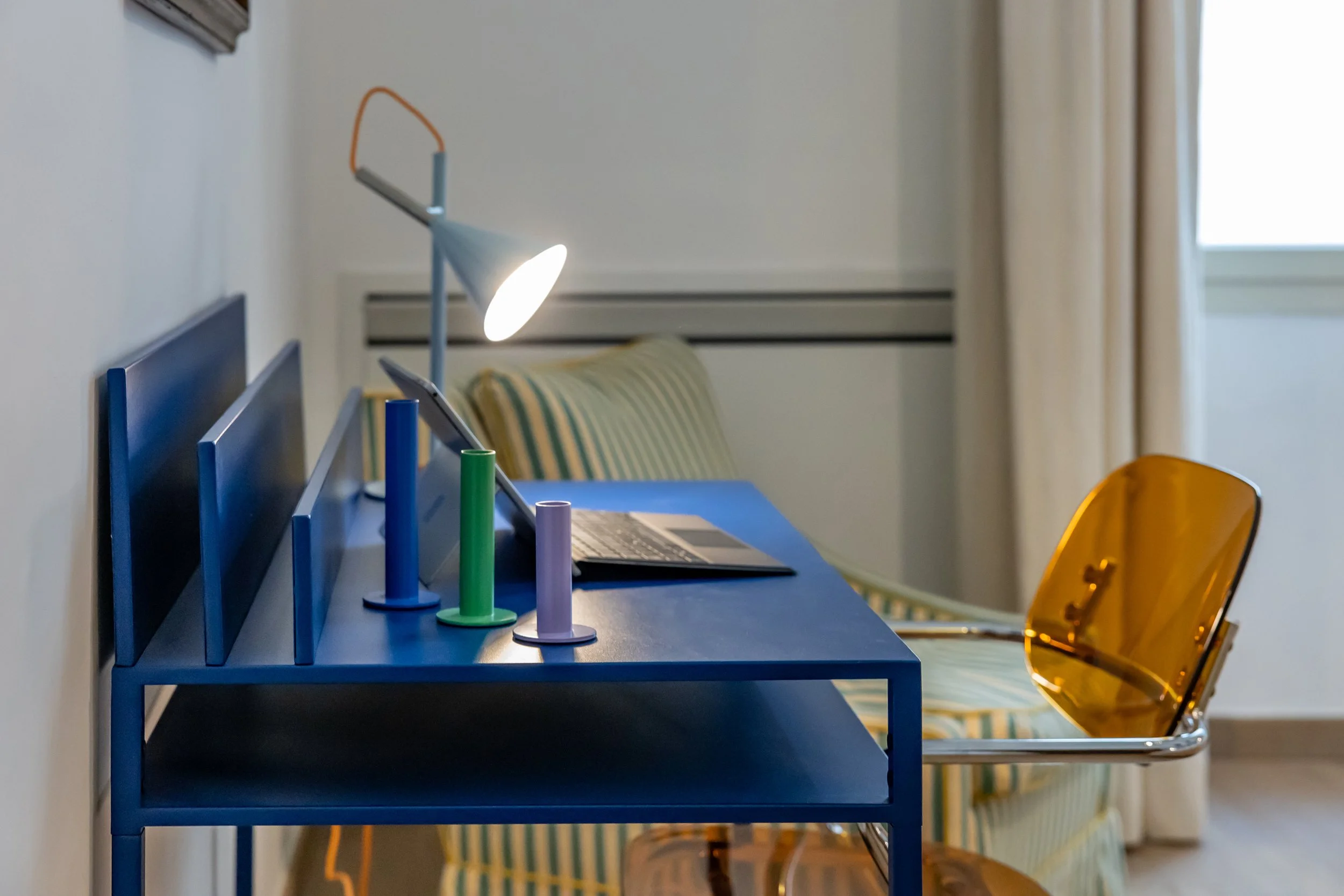 A room with a blue desk holding four colored cans, a lit desk lamp, an open laptop, a chair with a transparent yellow backrest, a striped couch, and a window with beige curtains.