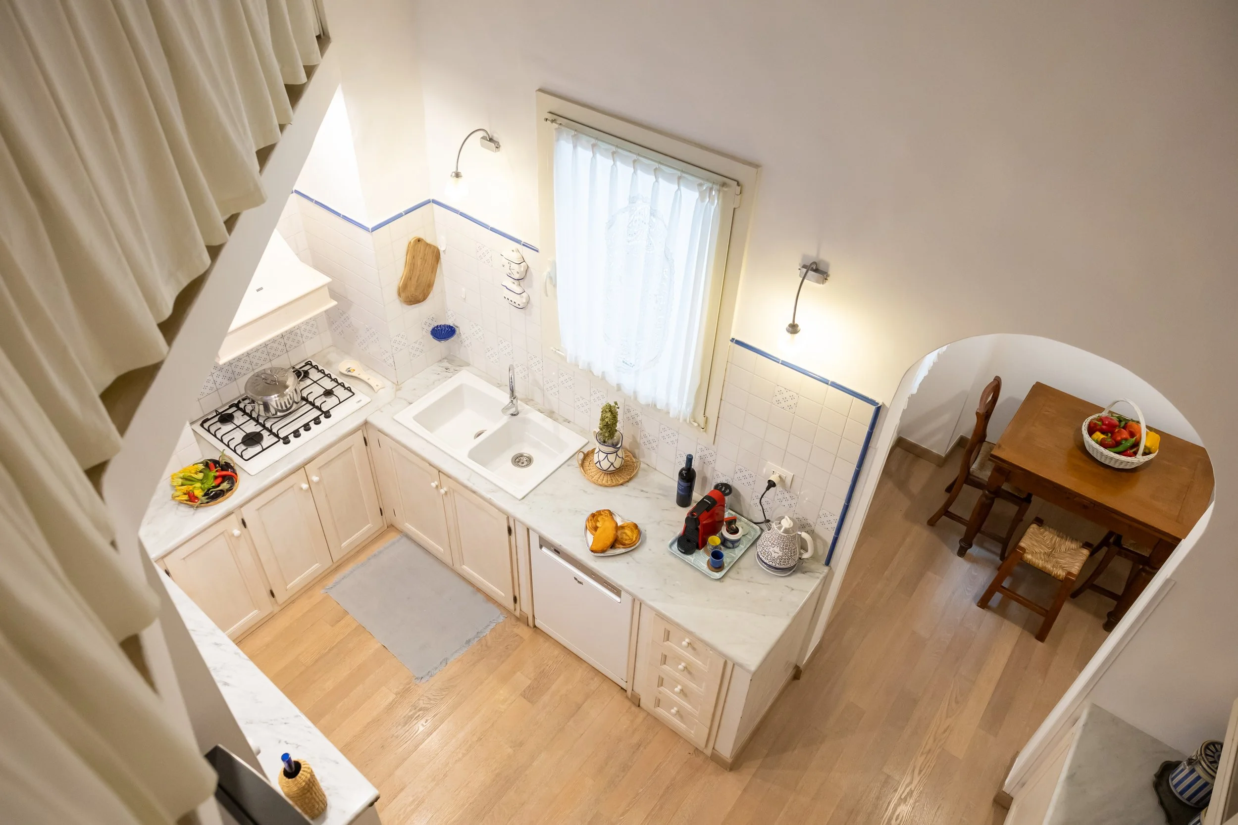 Overhead view of a kitchen with white cabinets, a marble countertop, and a double sink. There is a stove with a pot on it, a basket of vegetables, and various kitchen items on the counter. A window with white curtains is above the sink, and adjacent to the kitchen is a dining area with a wooden table and chairs, with a basket of fruit on the table.