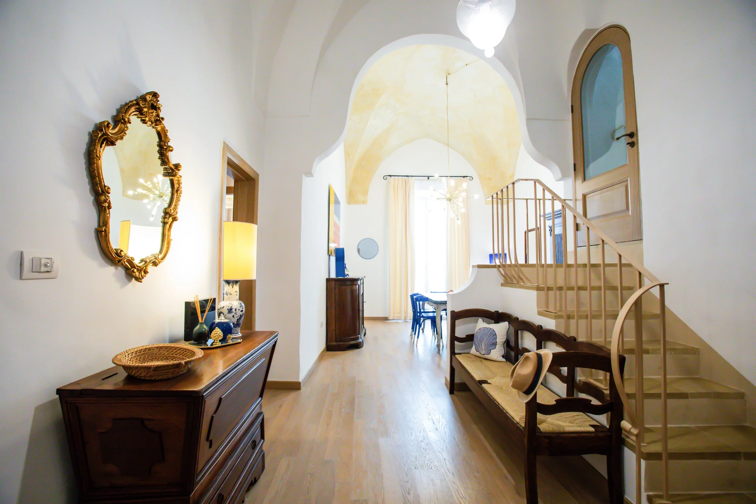 Bright living room with wooden flooring, a wooden sideboard, and a mirror on the wall. Further inside, there is a dining table with blue chairs near large windows with cream curtains. The room features an arched ceiling painted in a warm yellow hue and a staircase with a wooden railing leading up to the second floor.