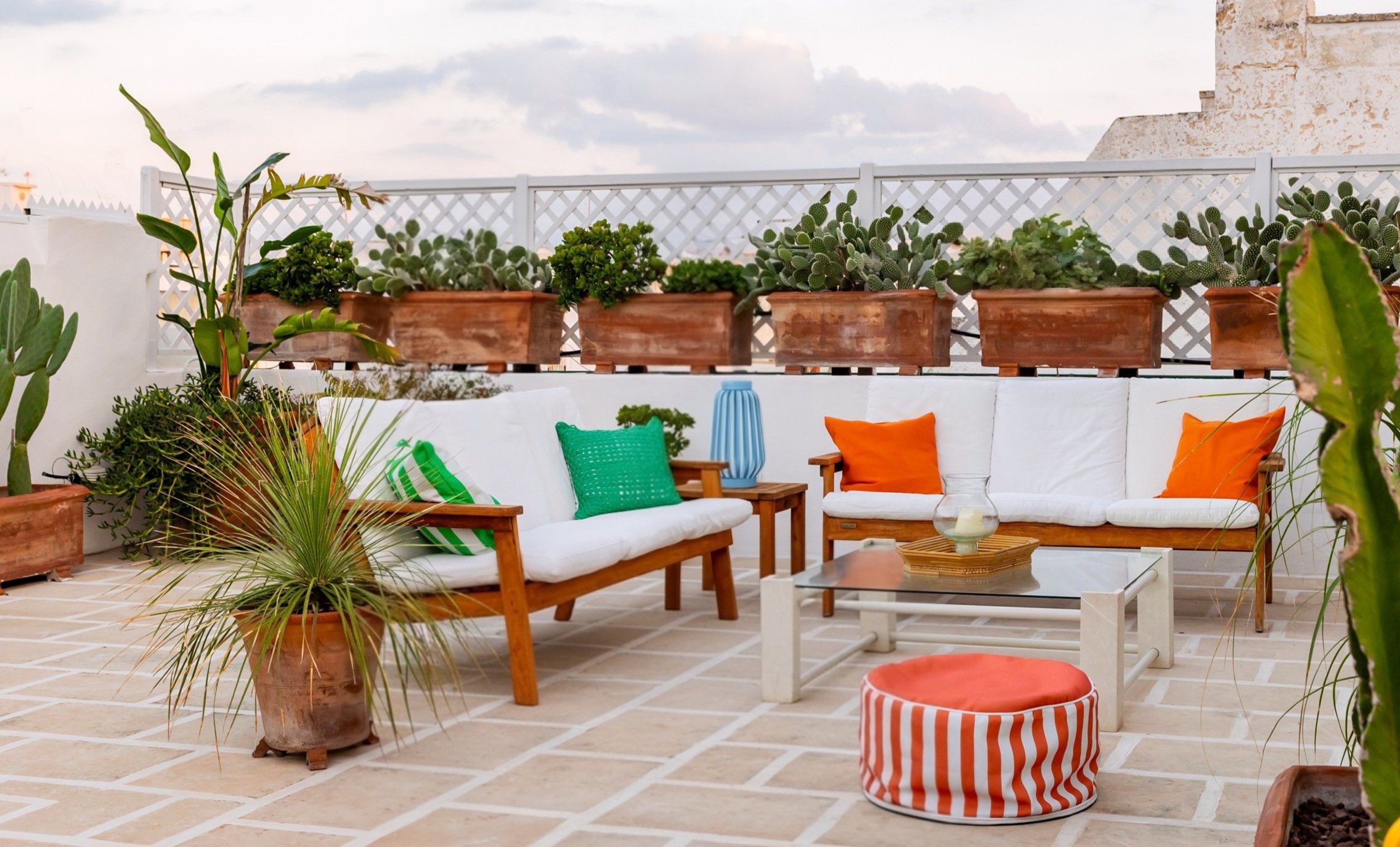 A rooftop patio with white cushioned wooden chairs and a glass coffee table, decorated with colorful pillows, surrounded by potted plants and succulents against a sky with clouds.