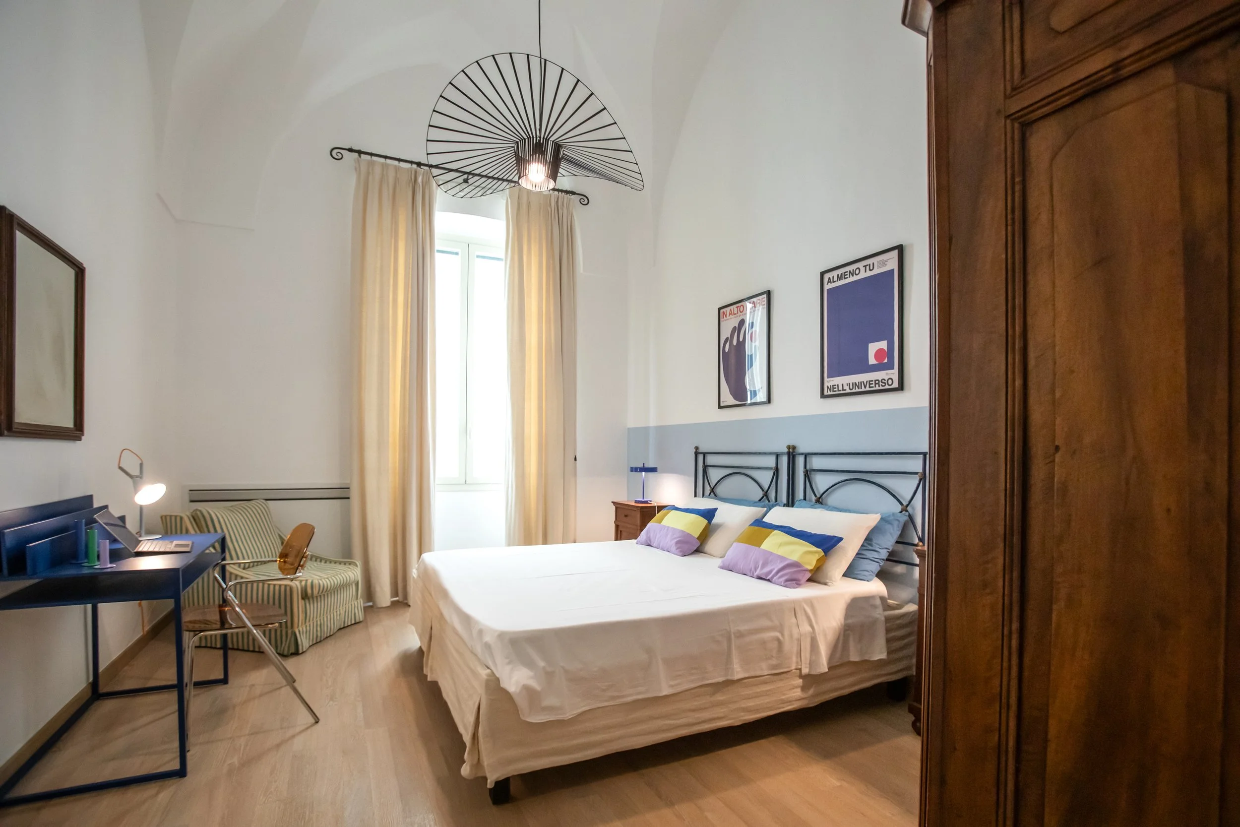 A bedroom with white walls, a large window with cream curtains, a metal bed frame with white bedding and colorful pillows, a wooden wardrobe, a desk with a lamp and books, an armchair with green and white stripes, framed art on the wall, and a modern ceiling light fixture.