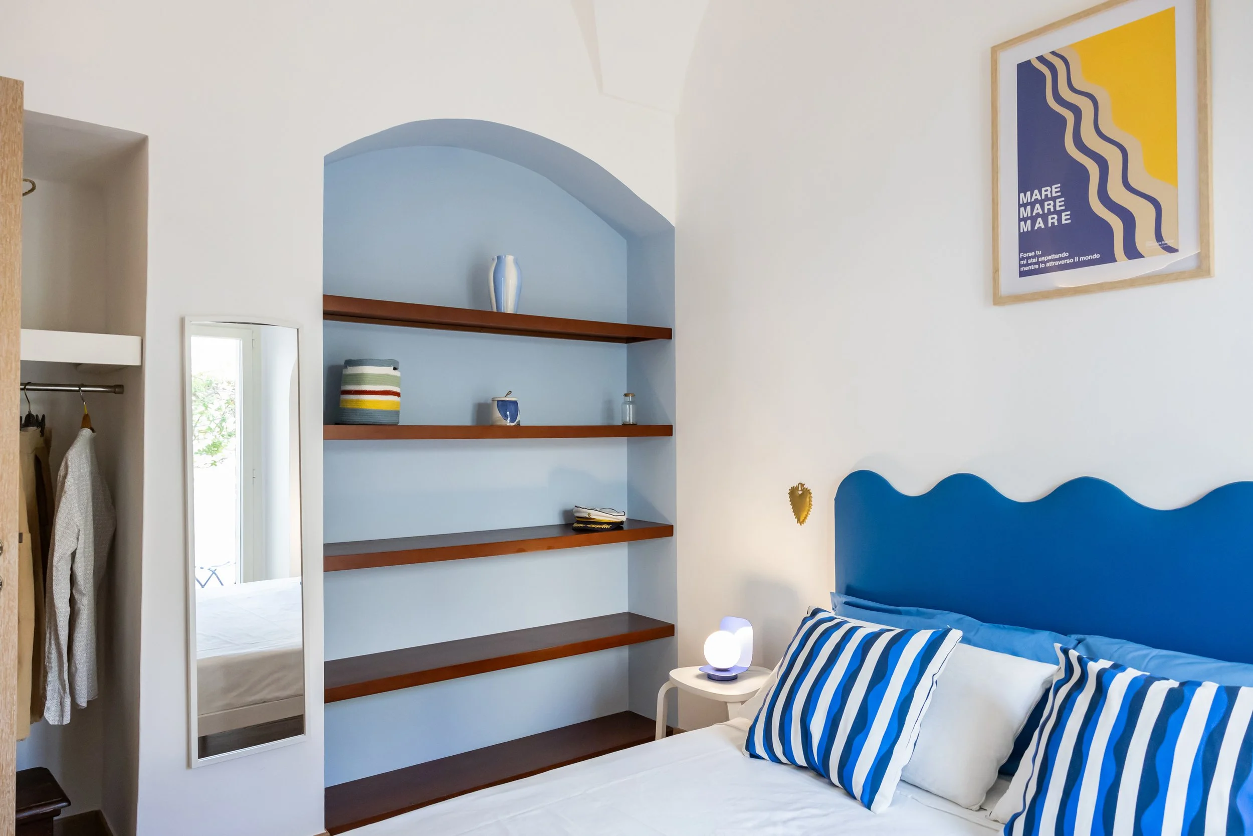 Bedroom with a blue bed headboard, white walls, striped pillows, wall art, small bedside table with a spherical lamp, open wardrobe with hanging clothes, and built-in blue shelf with decorative items.