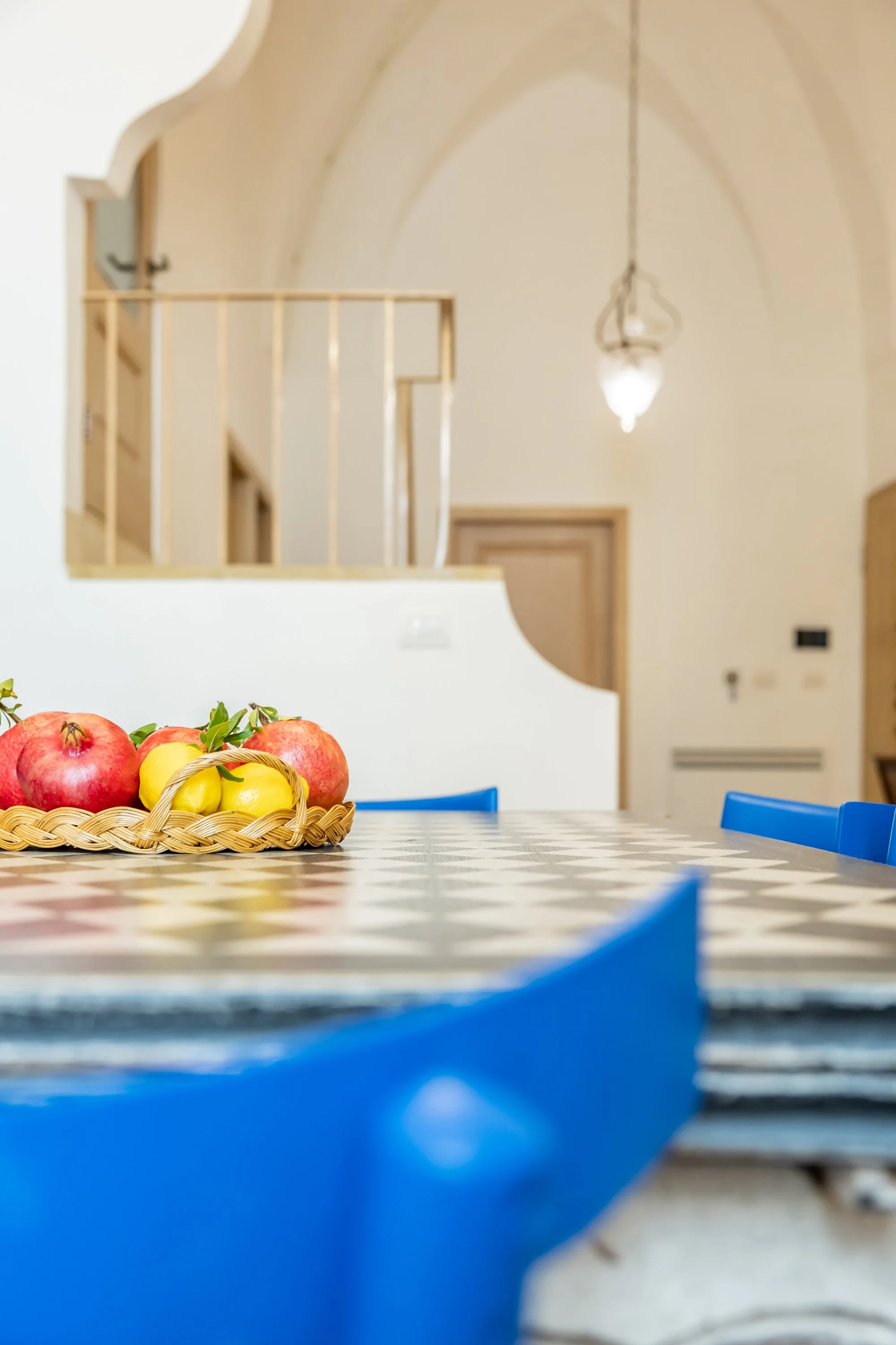 A basket of apples and yellow fruit on a checkered dining table with blue chairs, in a room with a vaulted ceiling and hanging light fixture.
