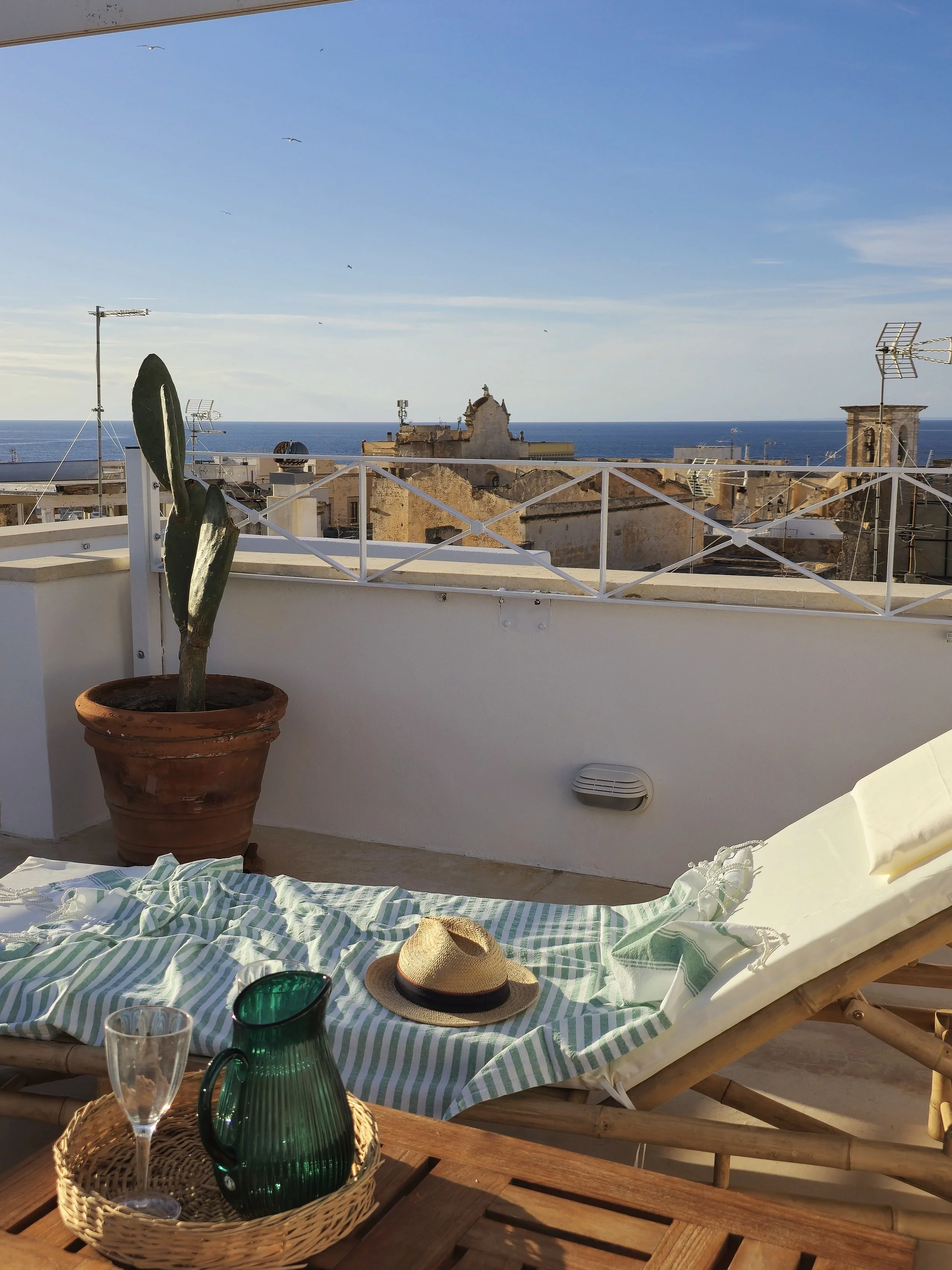 A rooftop terrace with a sun lounger, holiday hat, glass, and green pitcher on a basket, overlooking historic buildings and the sea in the background.