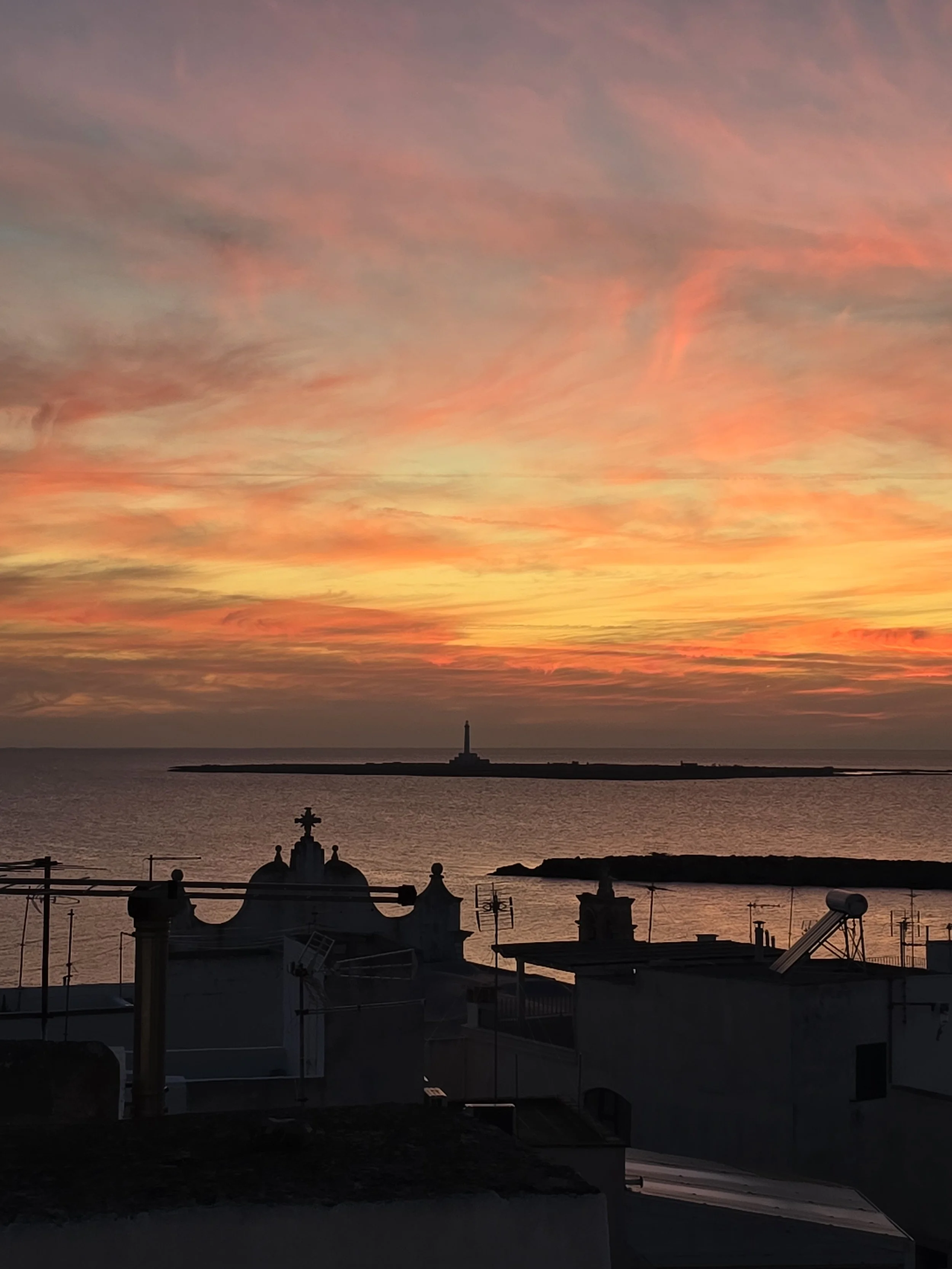 Panoramic rooftop at Palazzo Dolce Vita overlooking the historic rooftops and the sea, with the lighthouse of Sant’Andrea Island at sunset.