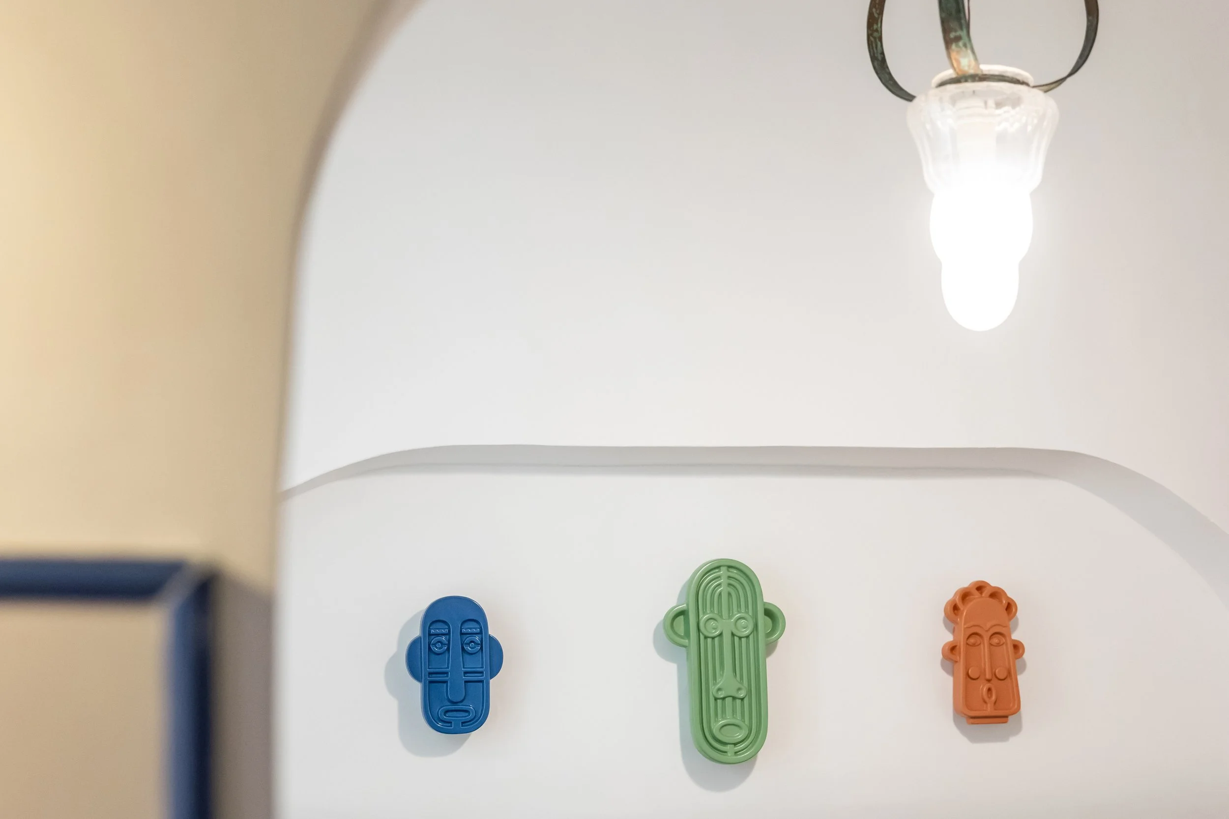Three colorful abstract face wall decorations in blue, green, and orange hanging on a white wall, with a ceiling light fixture visible at the top right.