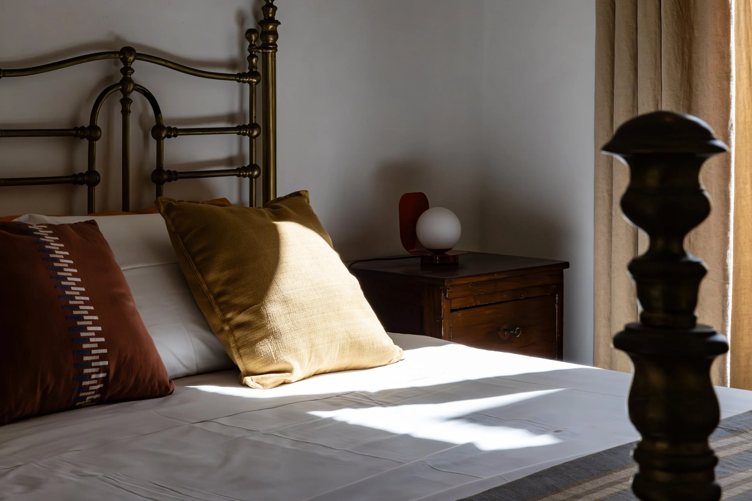 A neatly made bed with white sheets and several brown pillows, next to a dark wooden nightstand, a modern lamp, and beige curtains in a softly lit room.