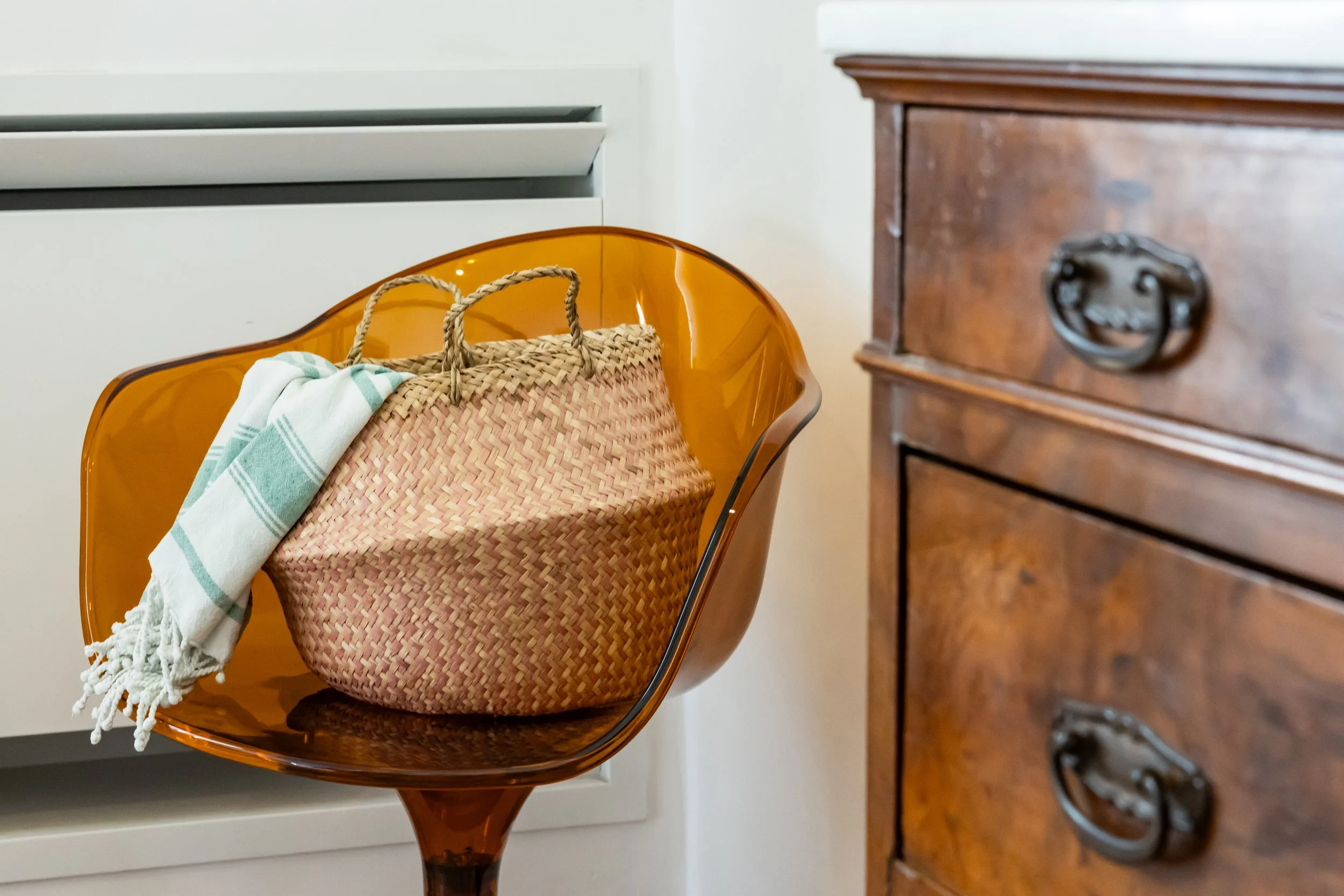 A vintage wooden dresser with ornate metal handles next to a modern amber-colored chair holding a woven straw bag and a patterned cloth.