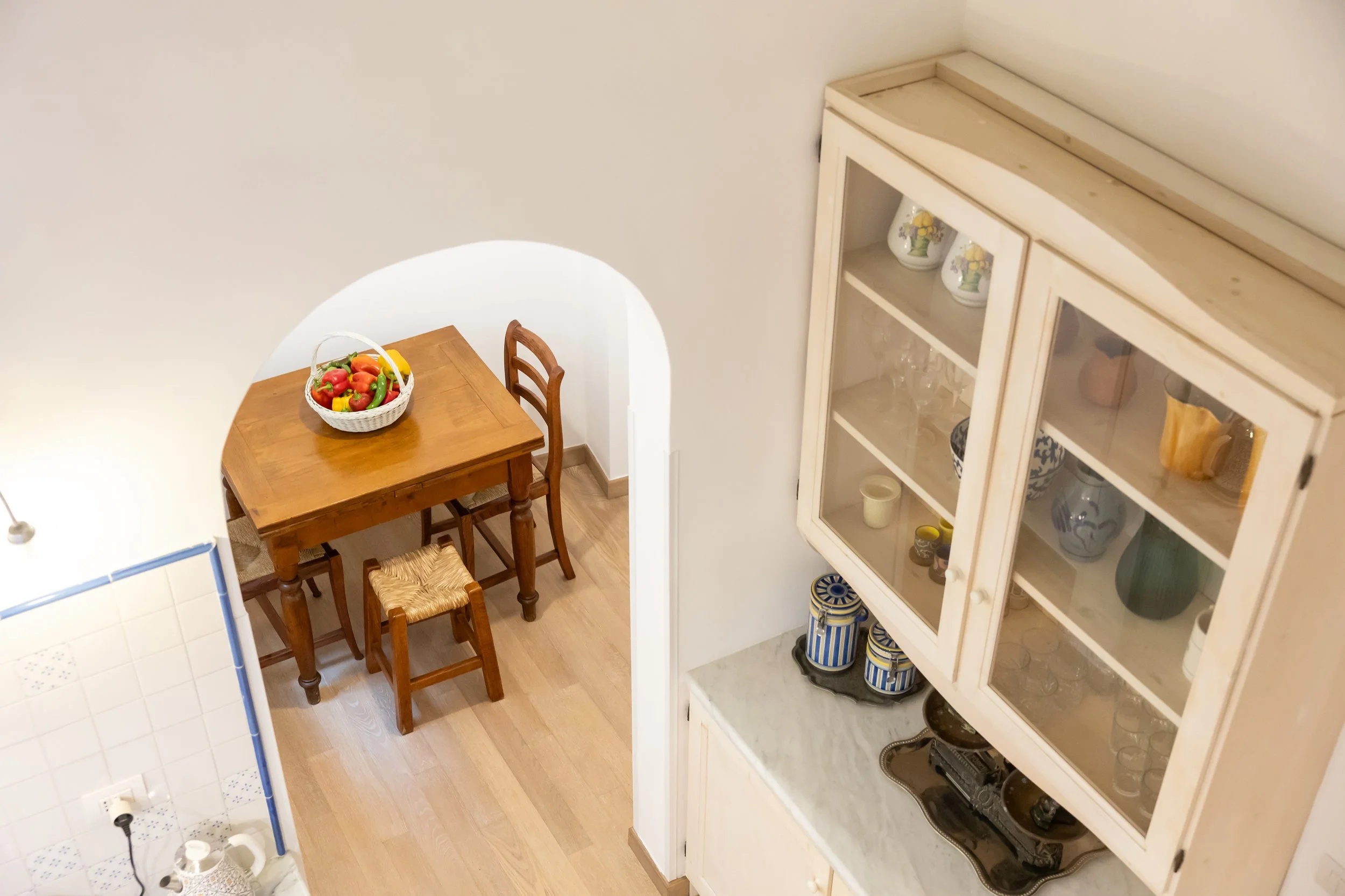 A cozy dining area with a wooden table and two chairs, a basket of colorful apples on top, next to a white cabinet with glass doors displaying various dishes and glassware.