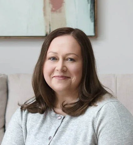 A white woman in her early-forties, with blue eyes and shoulder-length brown hair, wearing a light grey cardigan with silver buttons. She is smiling while sitting on a beige couch, with modern abstract art hanging on the wall behind her.