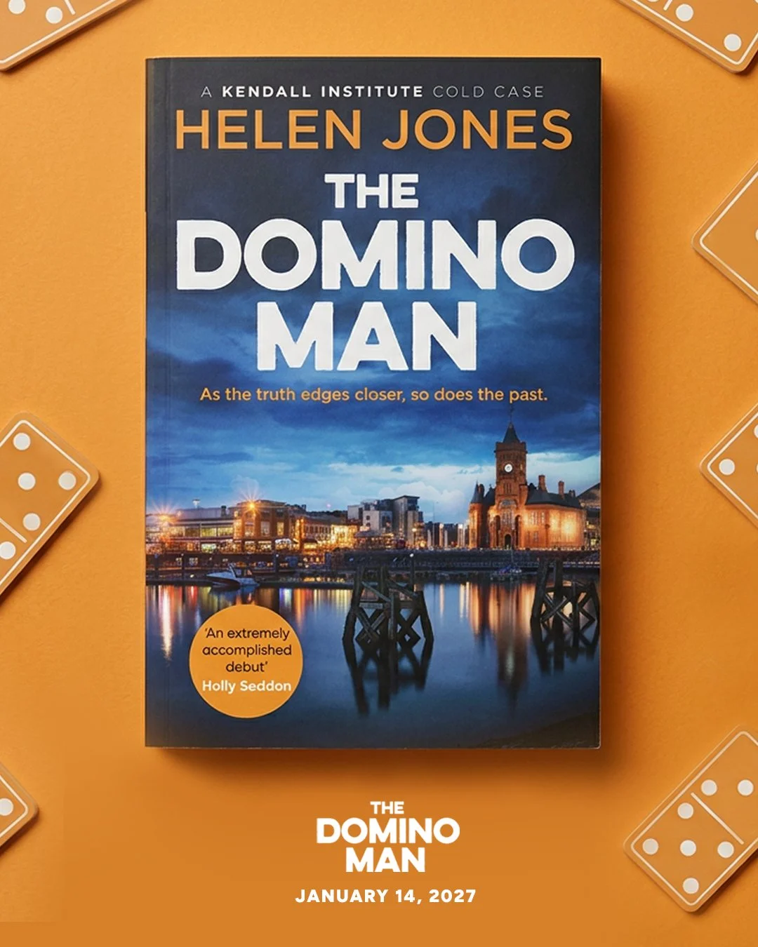 Book titled 'The Domino Man' by Helen Jones on an orange surface, with playing cards and dice around it. The cover features a night cityscape with a river, reflections, and a clock tower.