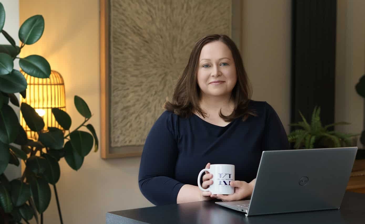 A white woman in her early-forties, with blue eyes and shoulder-length brown hair. She is sitting at a black table using a laptop and holding a white coffee mug with a Kendall Institute logo. Background is a cozy room with plants and wall art.