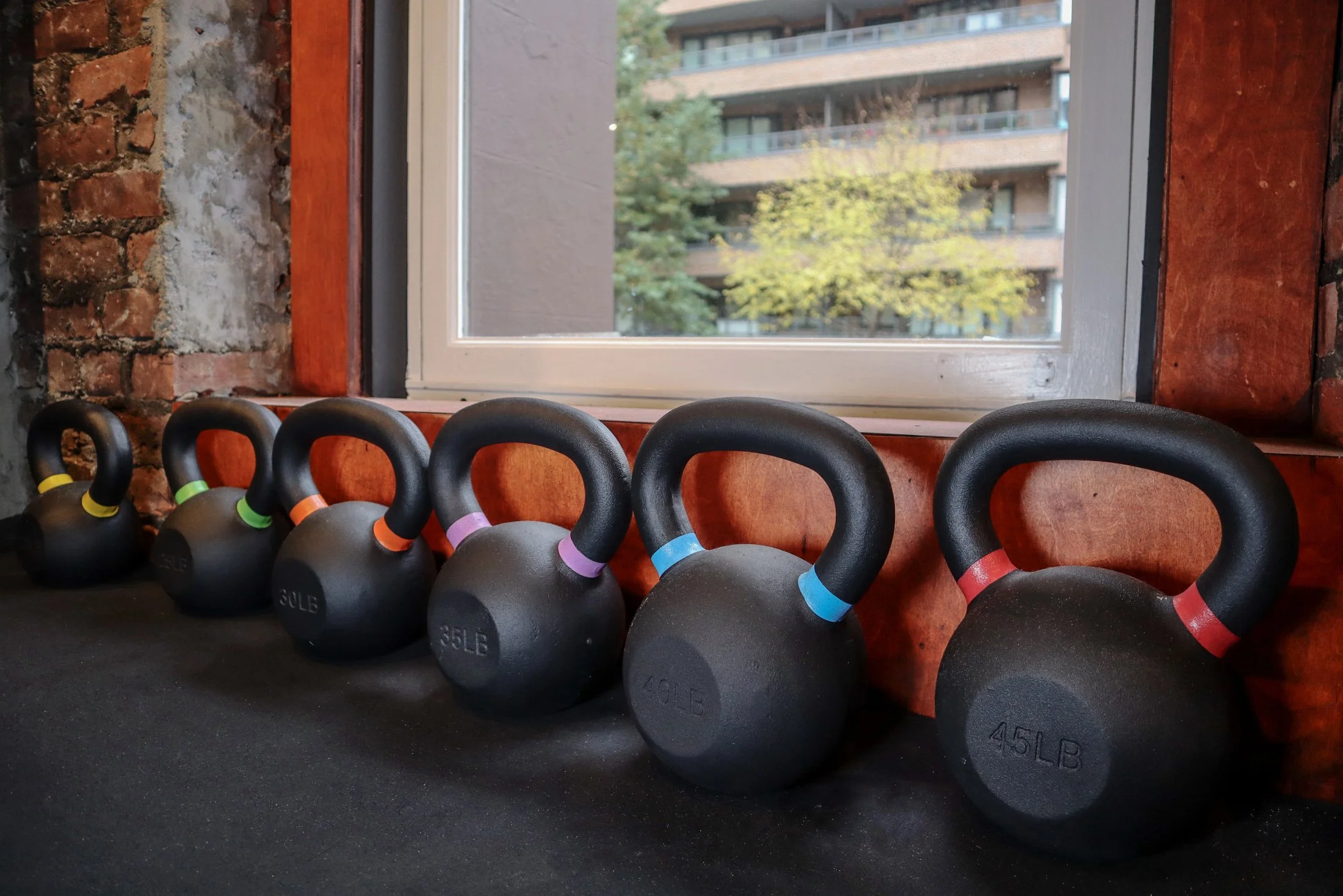 Kettlebells with different weights lined up on a black mat next to a window with a brick wall background.