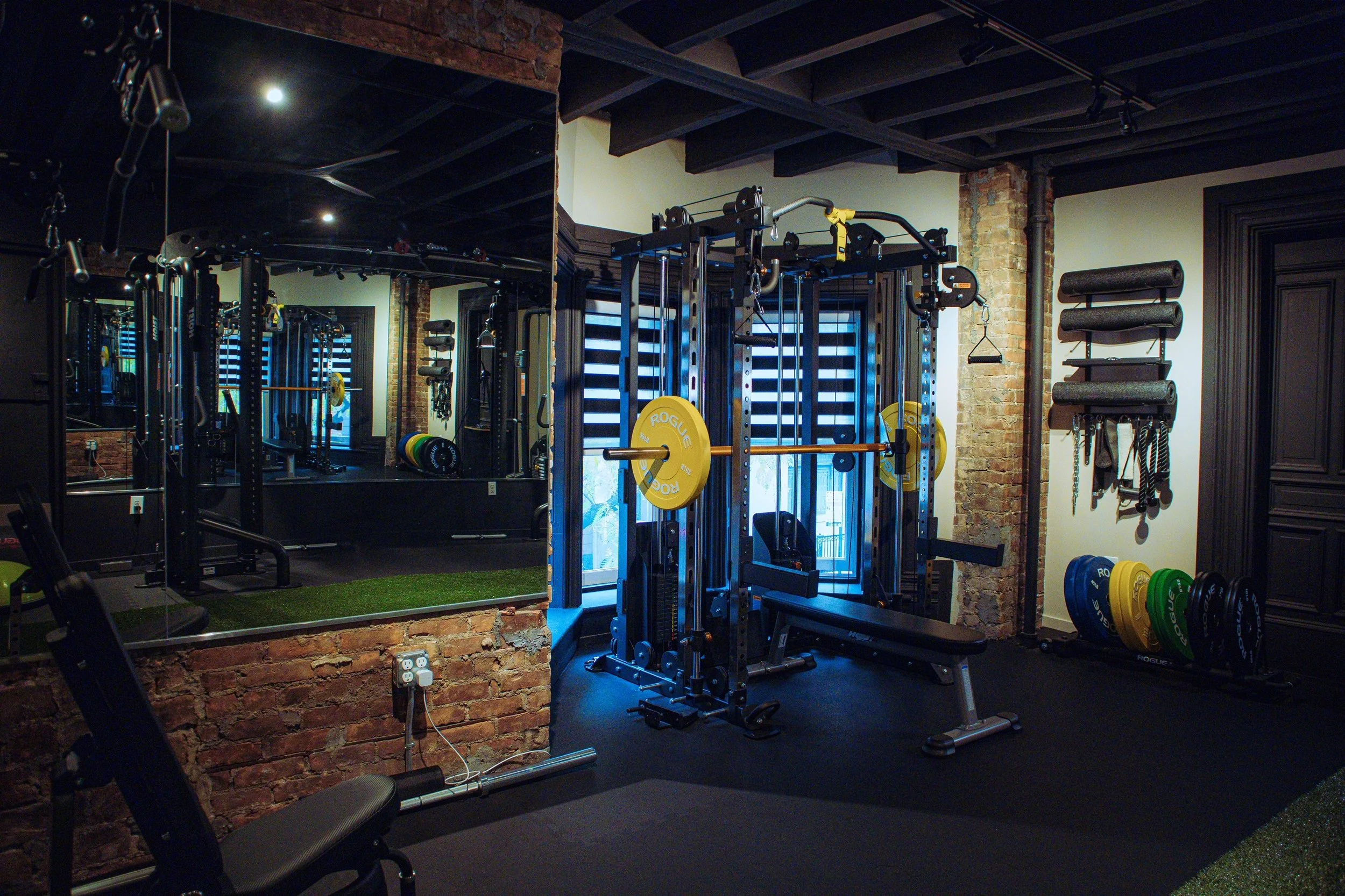 A modern gym with black walls and exposed brick, featuring gym equipment including weight plates, a squat rack, and a bench press, with large mirrors and window blinds.