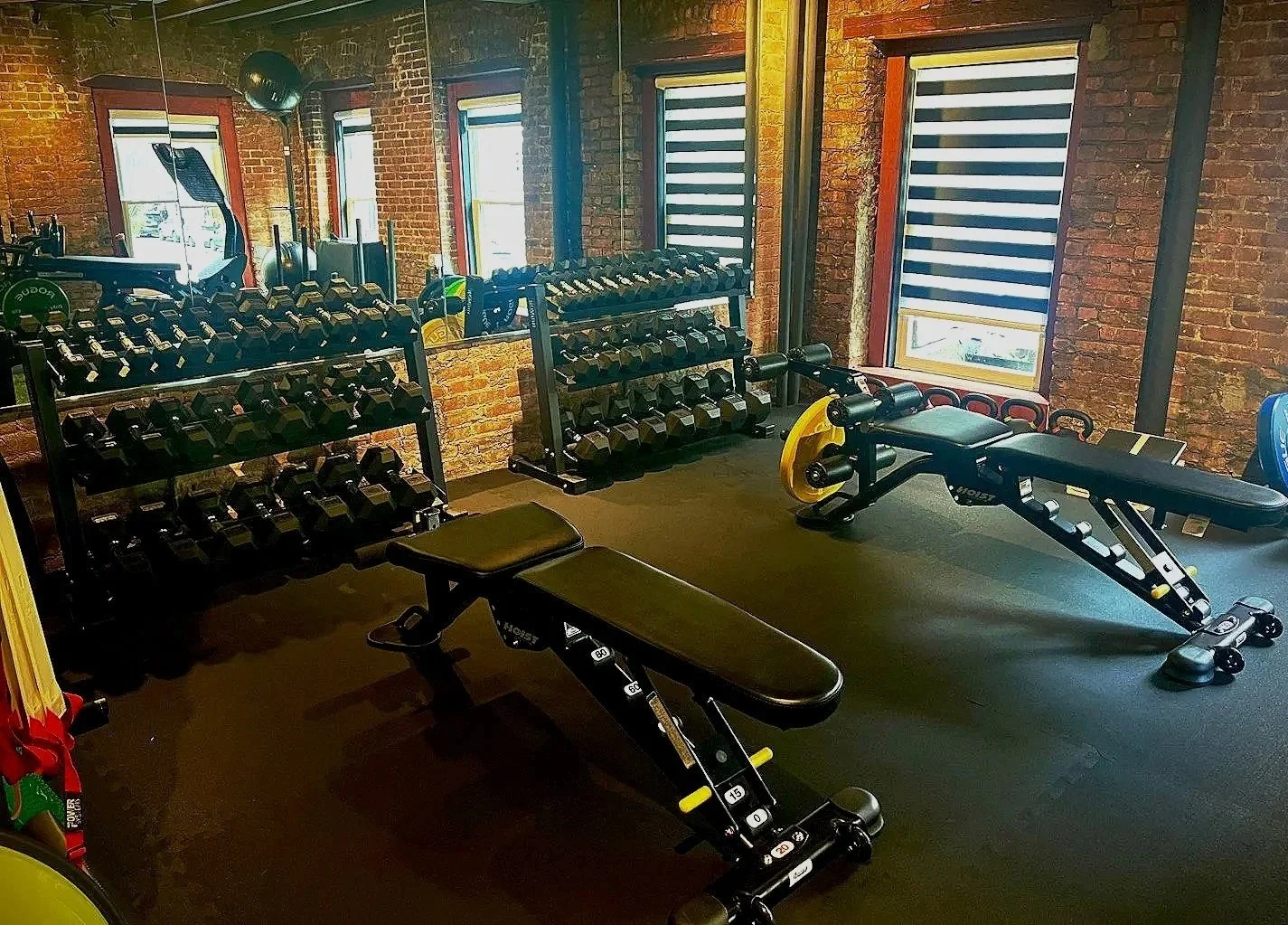 Interior of a gym with brick walls, black rubber flooring, two adjustable workout benches, and two black dumbbell racks filled with dumbbells. There are large windows with striped blinds allowing natural light.