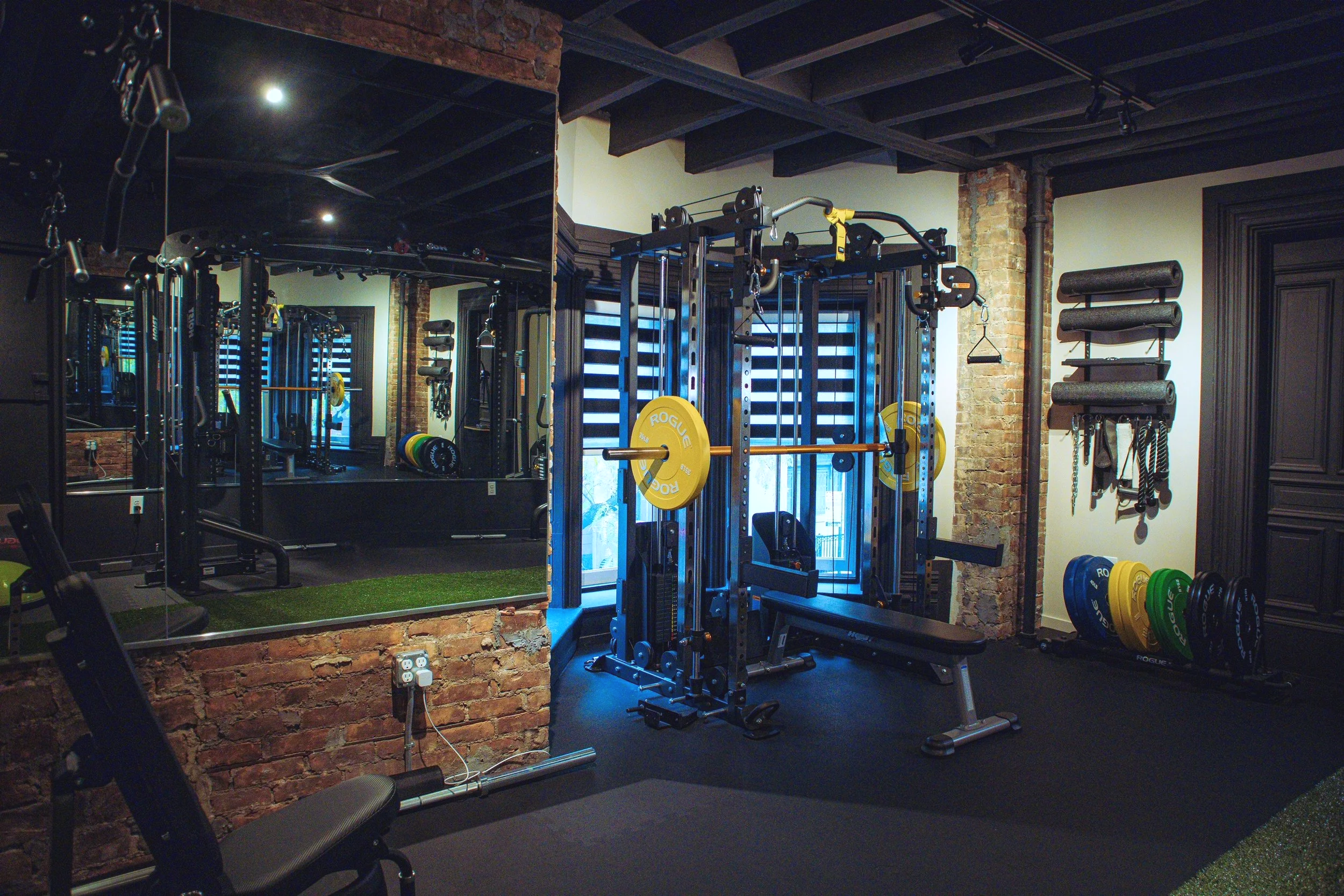 A gym with various workout equipment including a power rack with weights and exercise mats on shelves, with brick walls and mirrors.